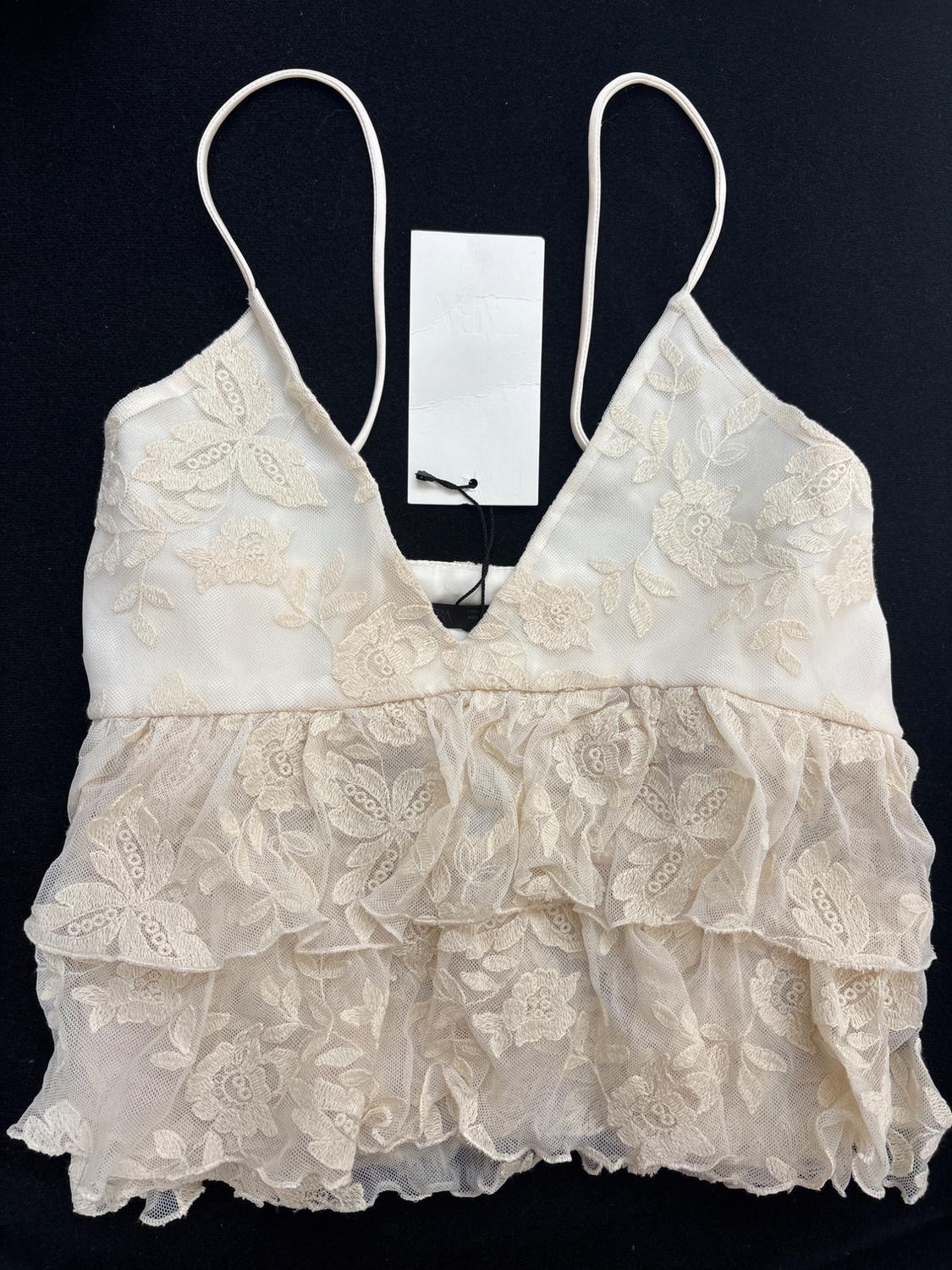 Cream camisole top with thin straps, V-neck, floral embroidery and lace frills, with a white label, on a plain black background.