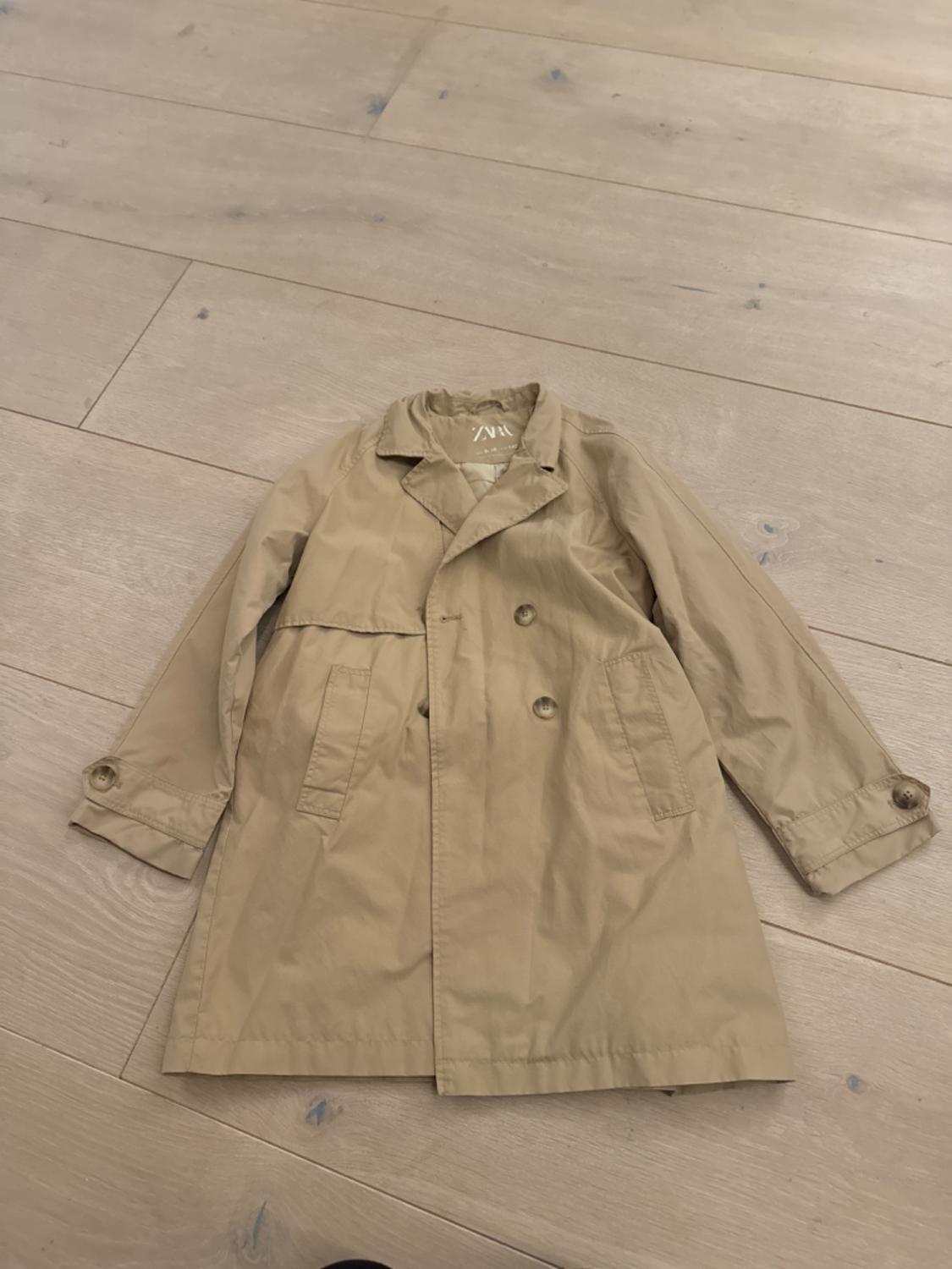 Double-breasted beige trench coat with front pockets, laid on a light wooden floor.