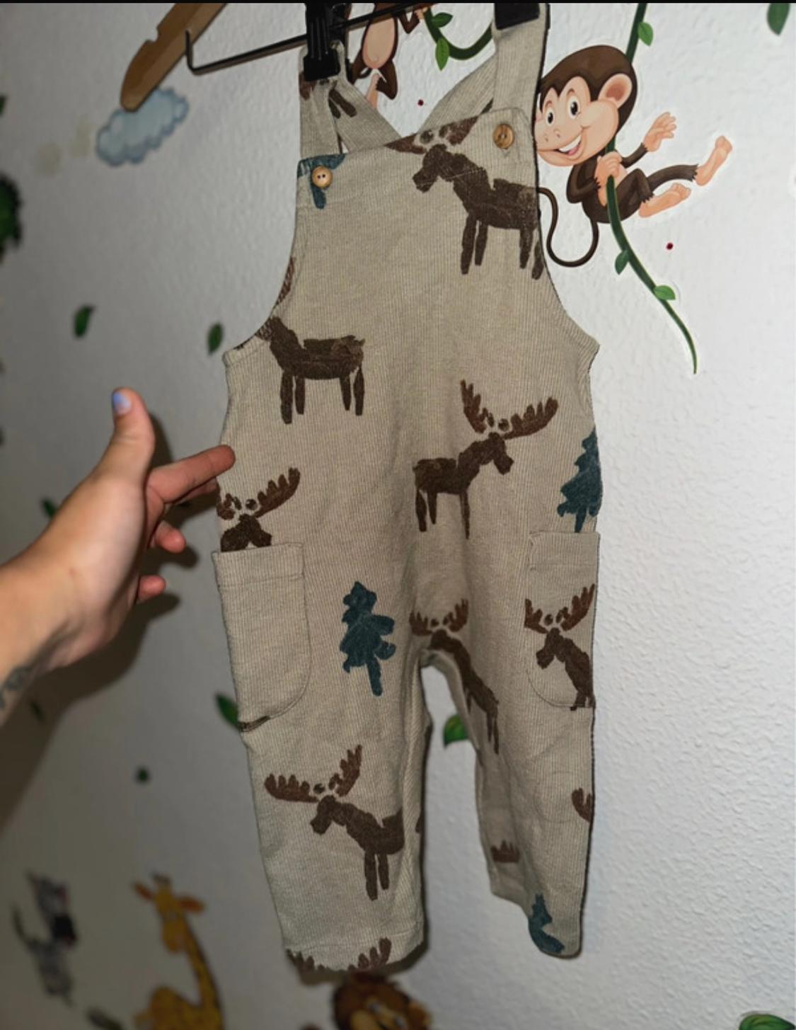 A beige children's all-in-one with a design of brown moose and green trees, hanging on a clothes hanger. The background shows drawings of animals and a hand.