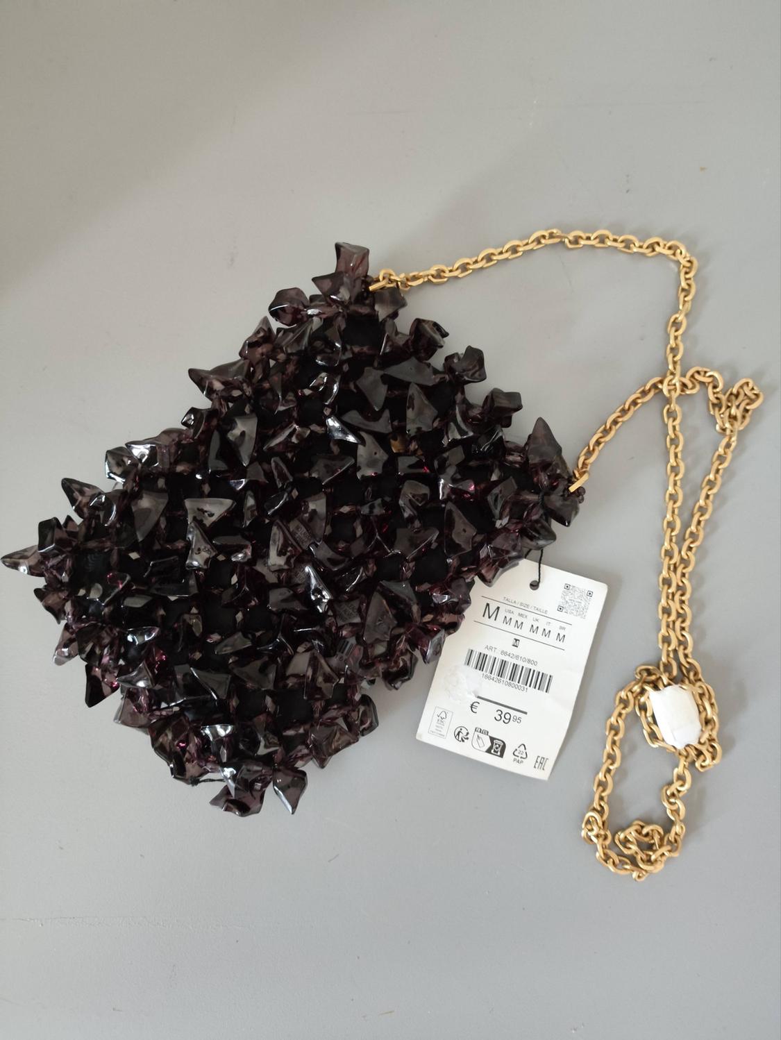 Dark purple bag with a spiked texture and gold chain. Tag with the text TALLA / SIZE / TAILLE M M M M M M € 39.95.