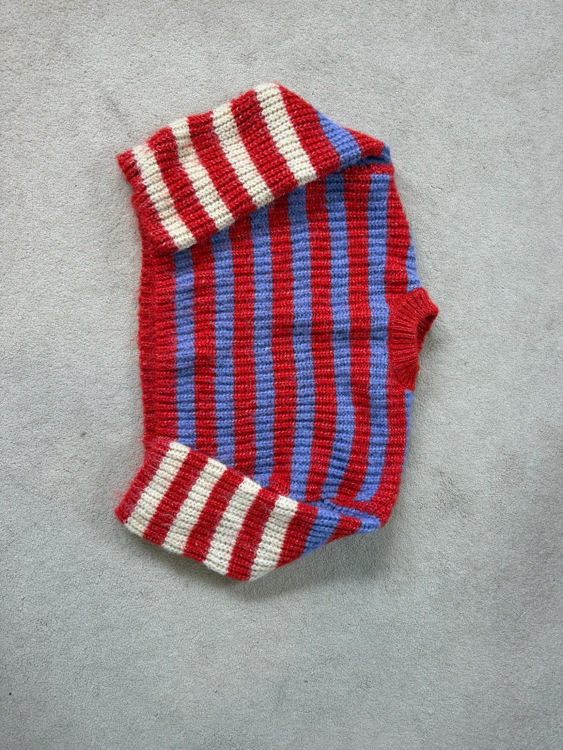 Folded knitted jumper against a grey background in a relaxed style. Red and blue striped body, with red and white striped sleeves.