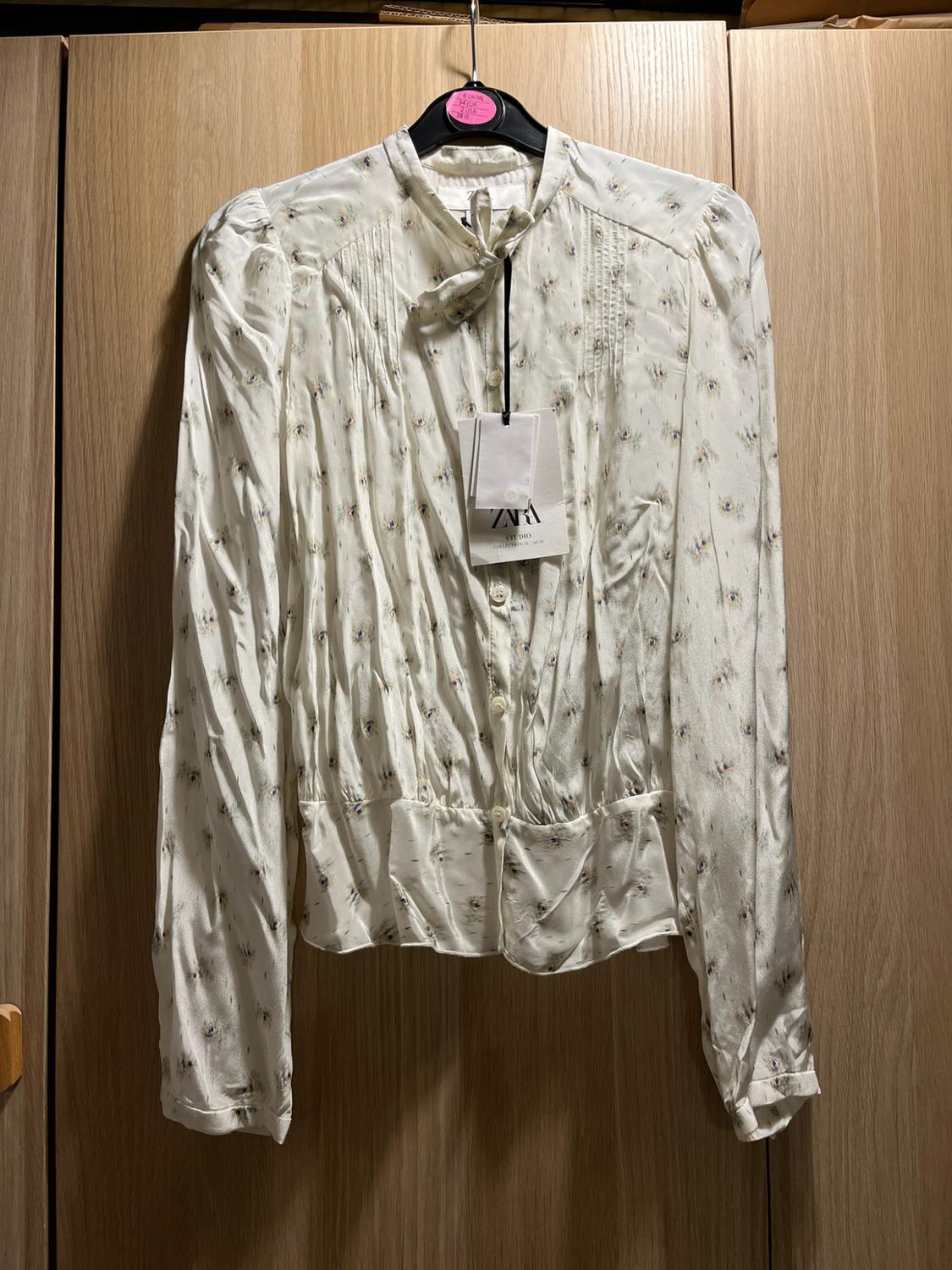 White blouse with subtle print, long sleeves, buttons and a bow at the neck, hanging in a wooden wardrobe. Label with text ZARA STUDIO.