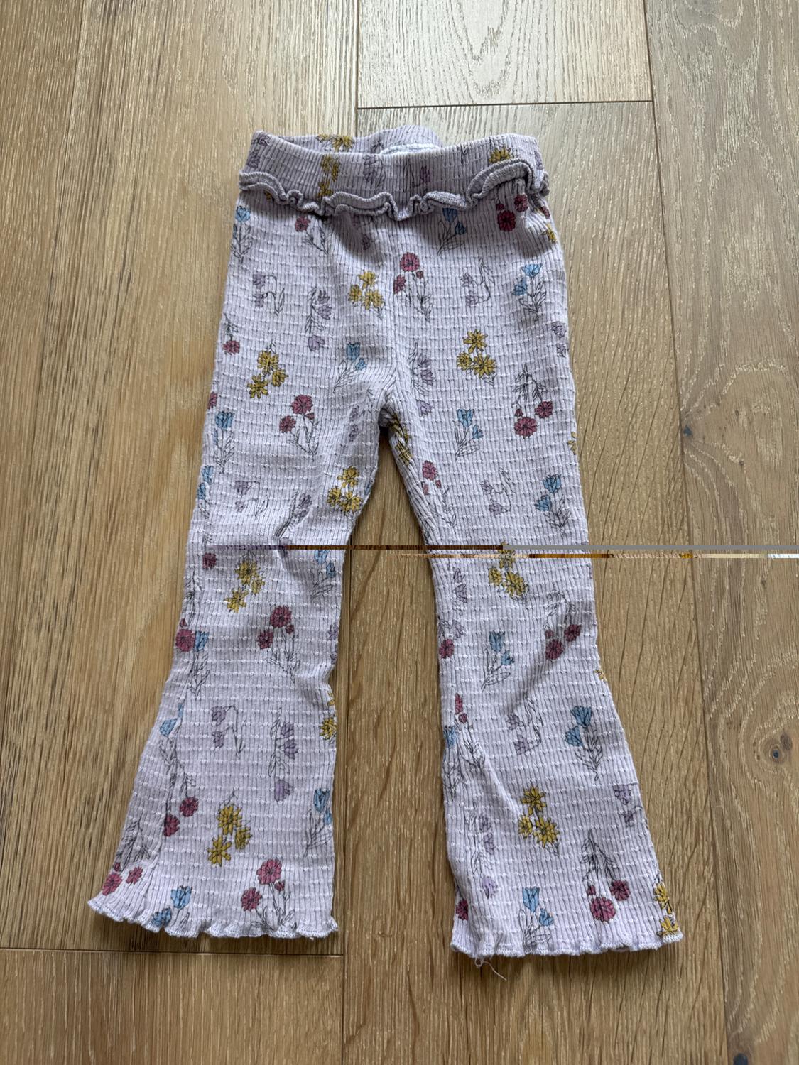 Children's crinkled fabric trousers with a floral print in yellow, red, and blue tones on a light background, on a wooden floor.