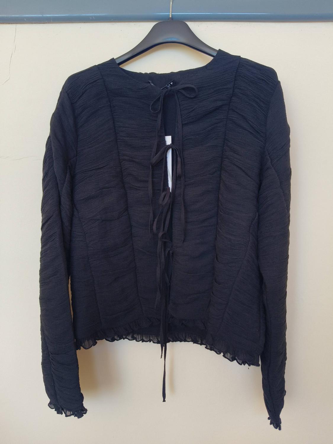 Black long-sleeved crinkled blouse, with a tie-front fastening and frilled edges, hanging on a hanger against a light-coloured wall.