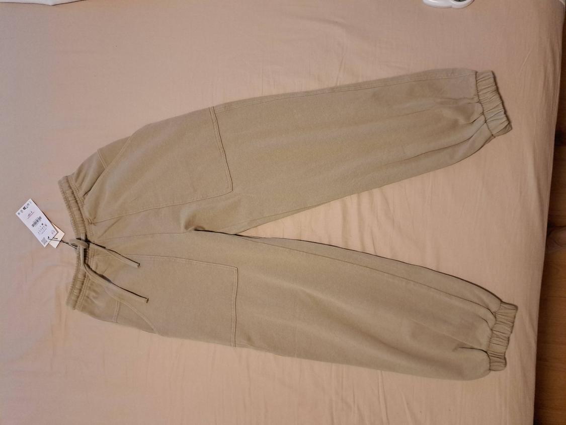Beige jogger trousers with tag, elasticated waist and cuffs, drawstring and pockets, laid out on a light-coloured casual-style sheet.