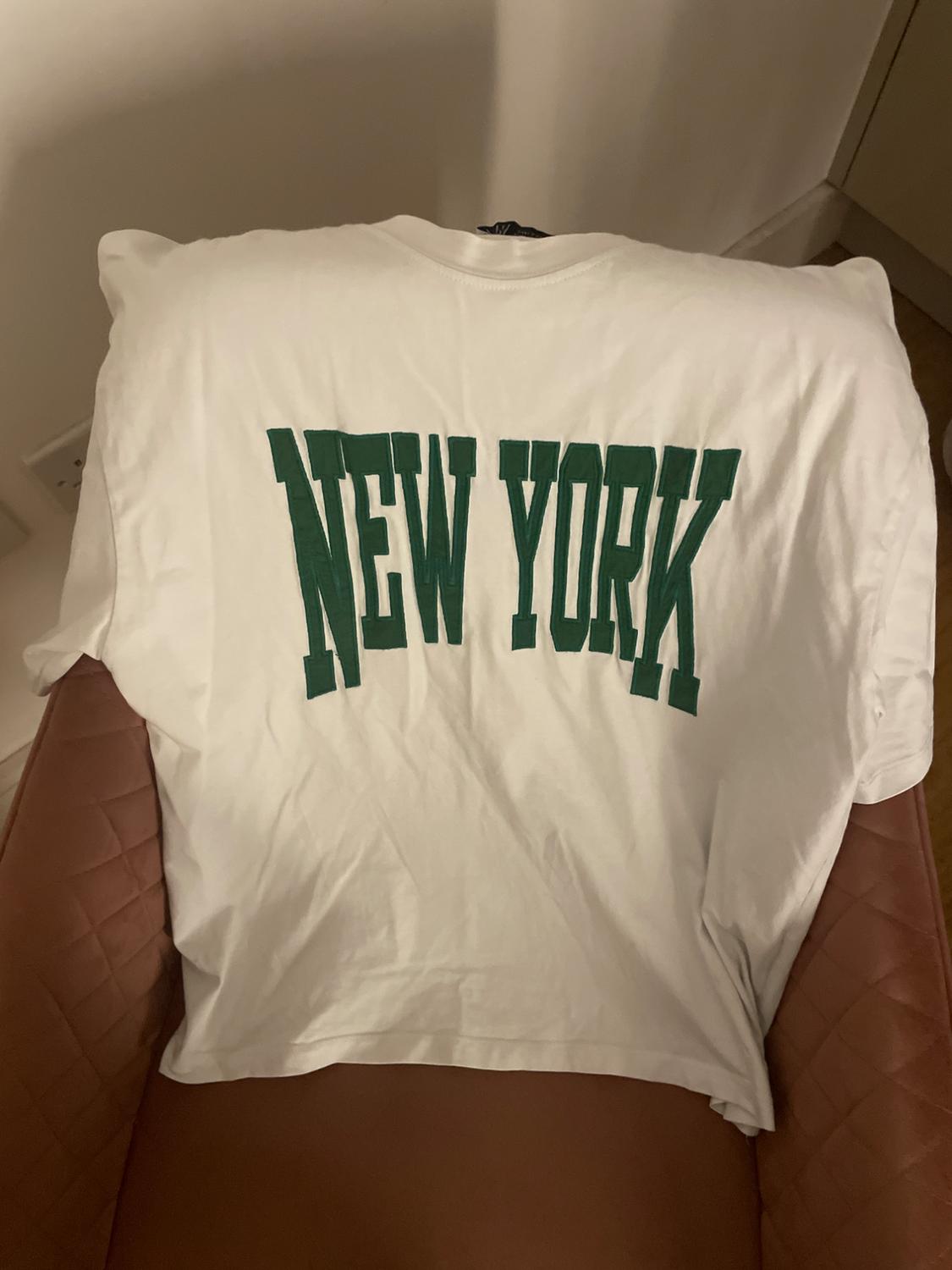 White T-shirt with "NEW YORK" text in green, placed on a brown chair in a brightly lit indoor setting.