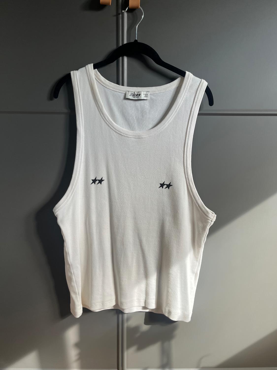 White sleeveless top with two black stars on the chest, hanging on a black hanger against a grey background.
