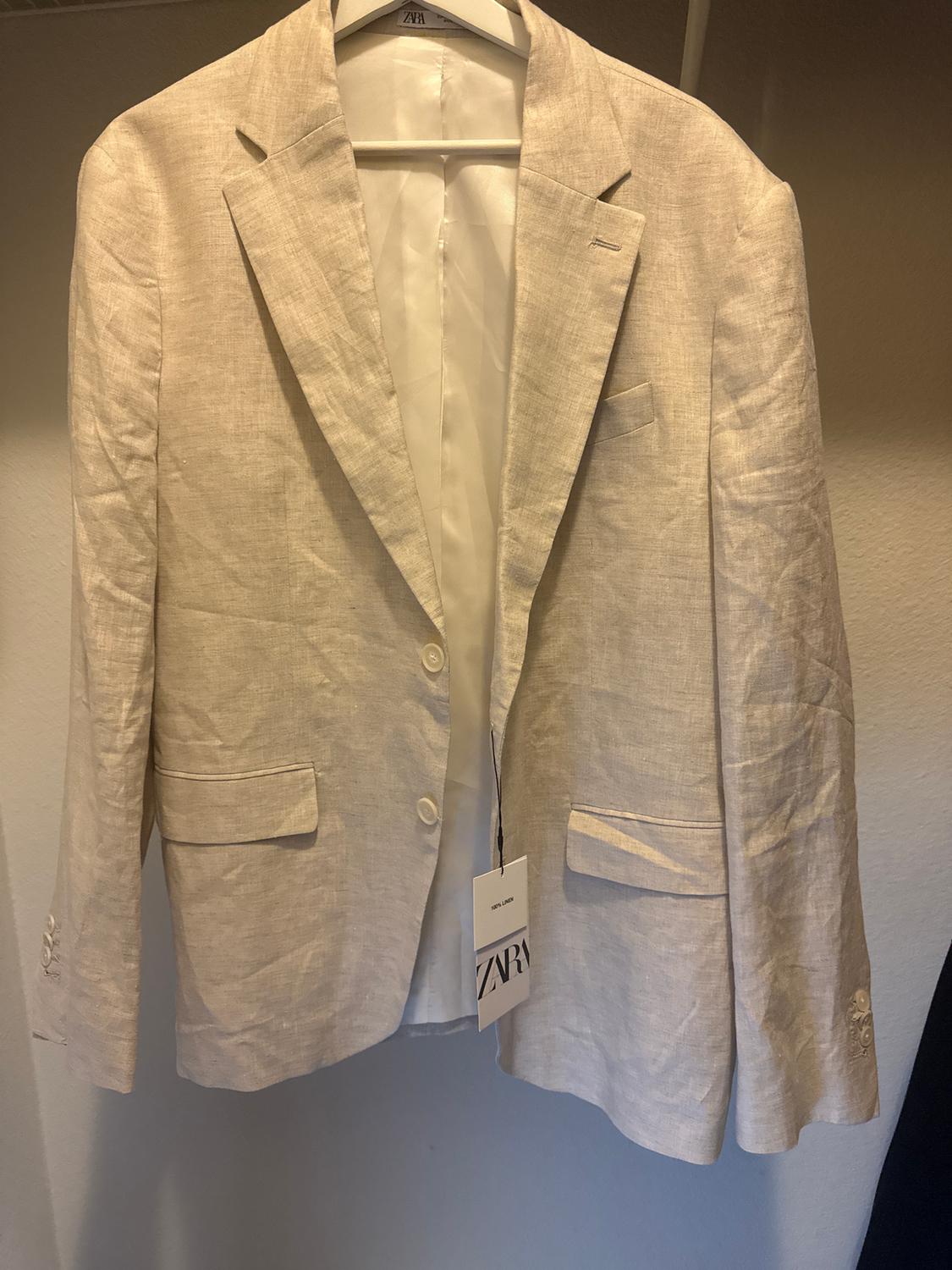 Beige linen jacket hanging on a hanger, with a visible "ZARA" label. Neutral background and soft lighting.