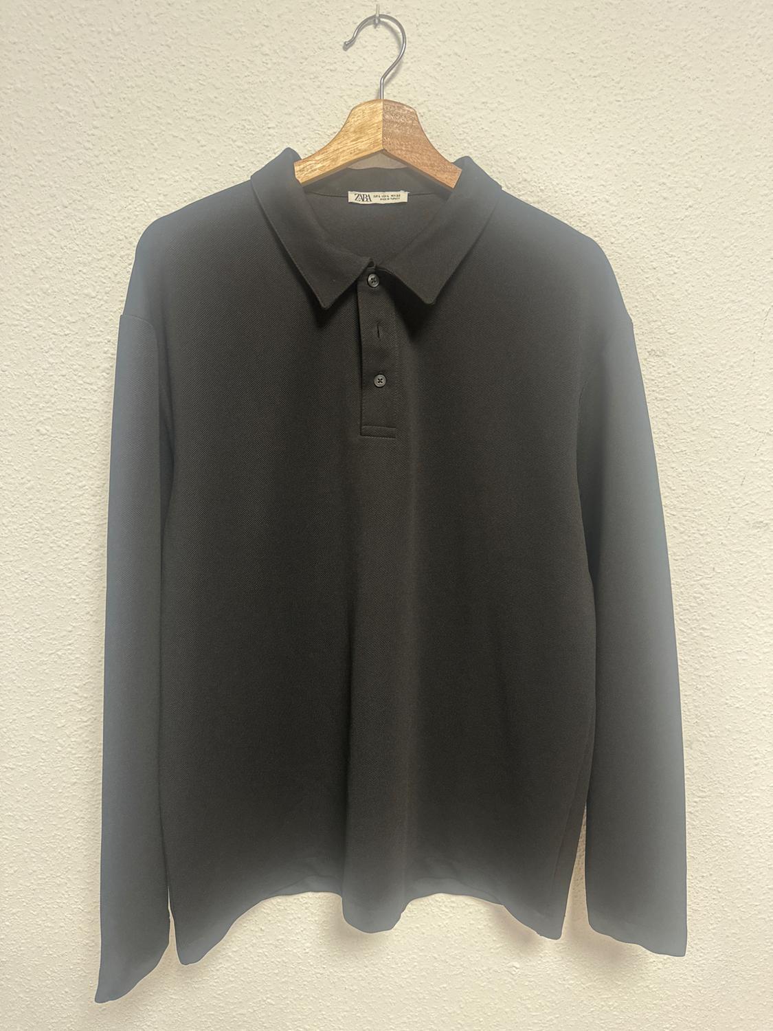 Black long-sleeved polo shirt with buttons, hanging on a wooden hanger against a white wall. Label visible.