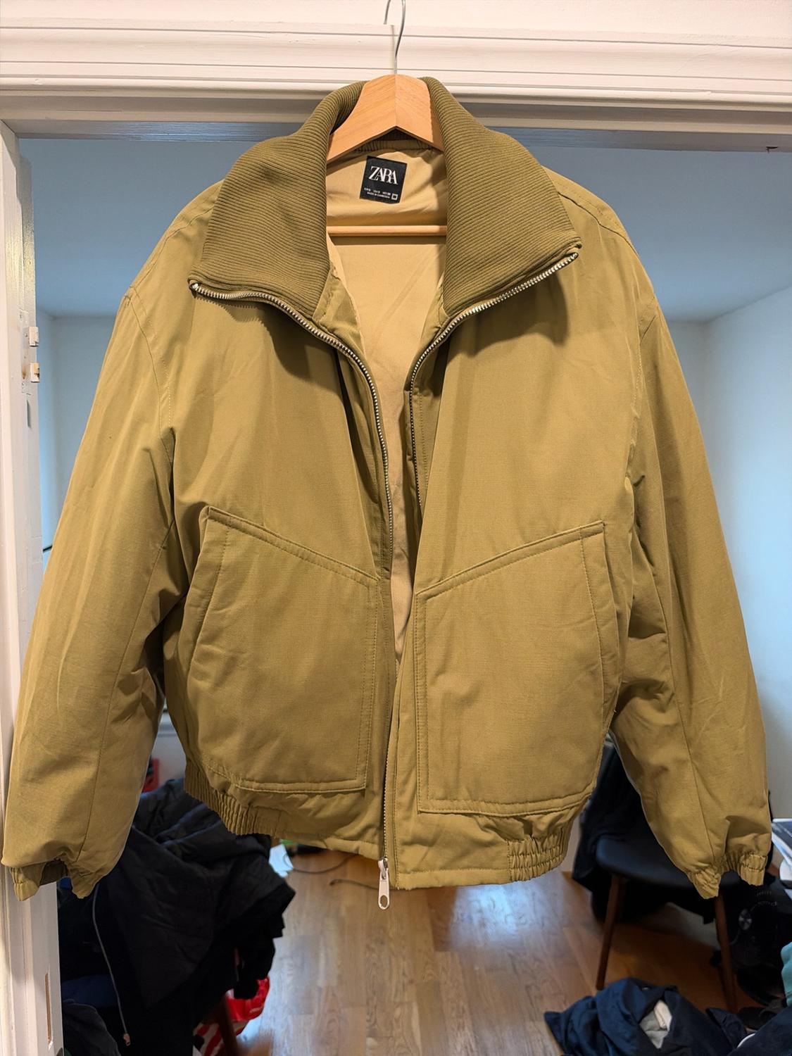 A khaki jacket with a zipper and pockets, hanging on a wooden hanger in a door frame. Interior label zara.
