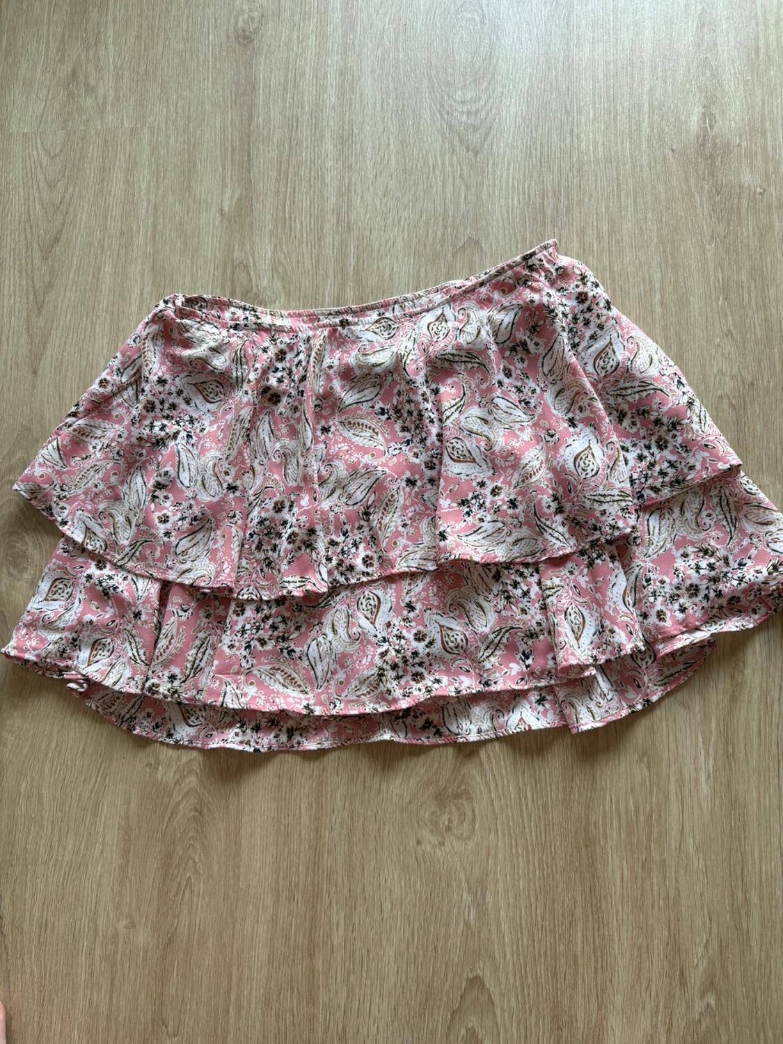 Short ruffled skirt with floral and paisley print in pink and white tones, on a light wooden floor.