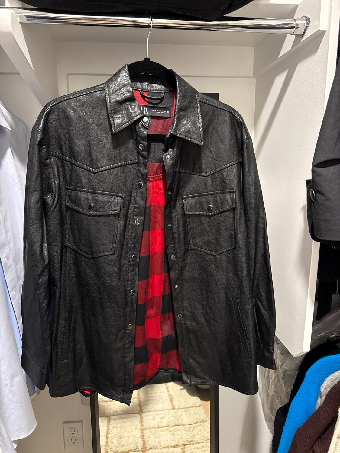 Black overshirt hanging in a white wardrobe. It highlights its red and black check lining, buttons, and front pockets. Text ZARA.