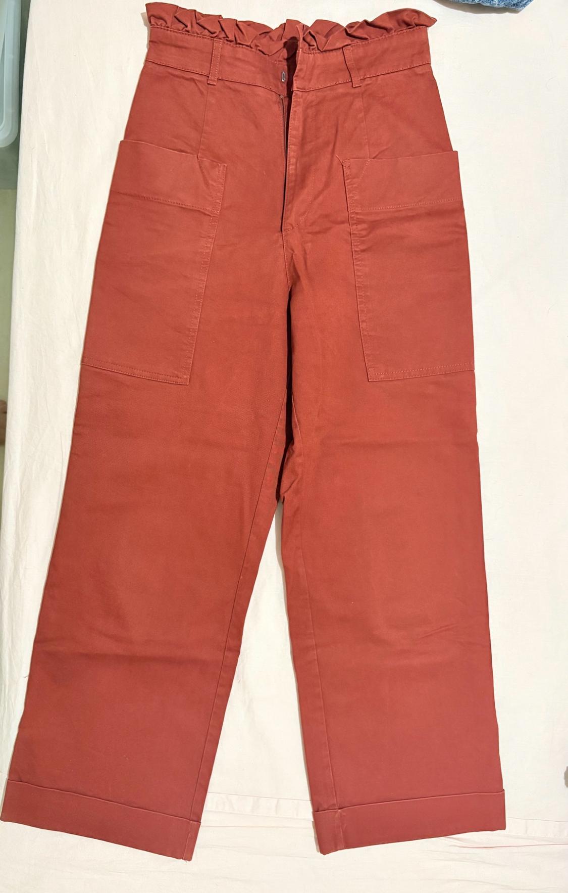 Rust-coloured, wide-leg, high-waisted trousers with a gathered waist, front patch pockets and turn-up hems, laid out on a white background.