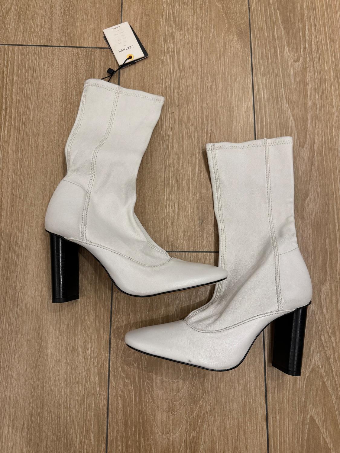 White high-heeled boots on a wooden floor. A tag with the word "LEATHER" hangs from one of the boots.