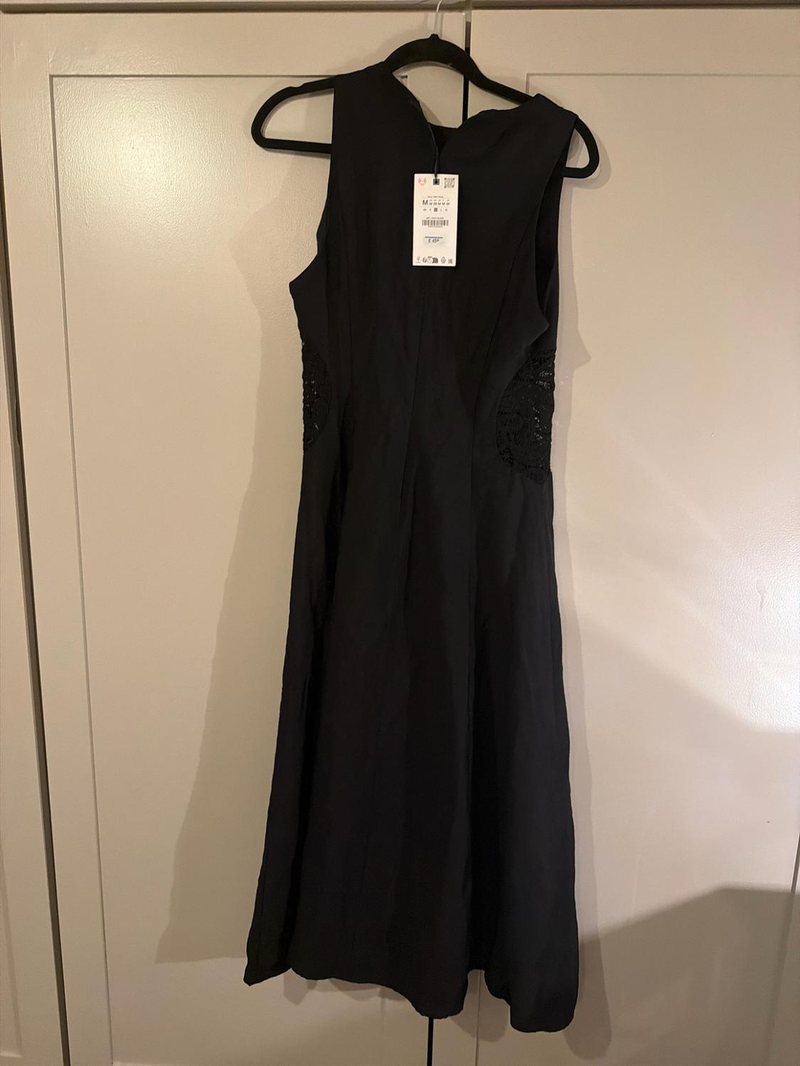 Sleeveless black dress with a label and lace detailing at the sides, hanging on a black hanger in front of a light-coloured wardrobe.