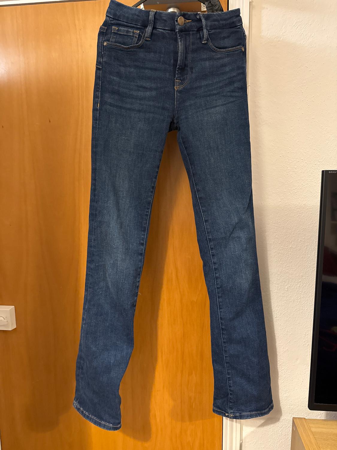 Blue jeans hanging on a coat hanger in front of a wooden door. Straight cut, five pockets and visible stitching.