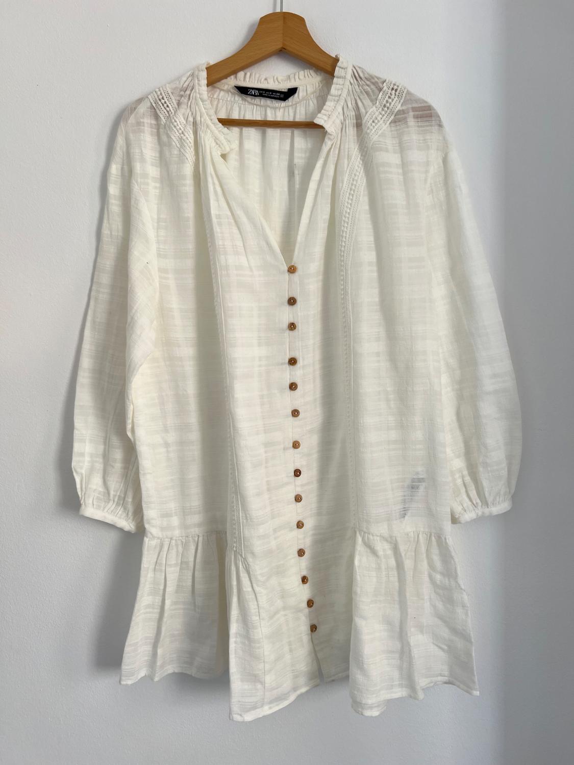 White, long-sleeved blouse with brown buttons, hanging on a wooden hanger against a white wall.