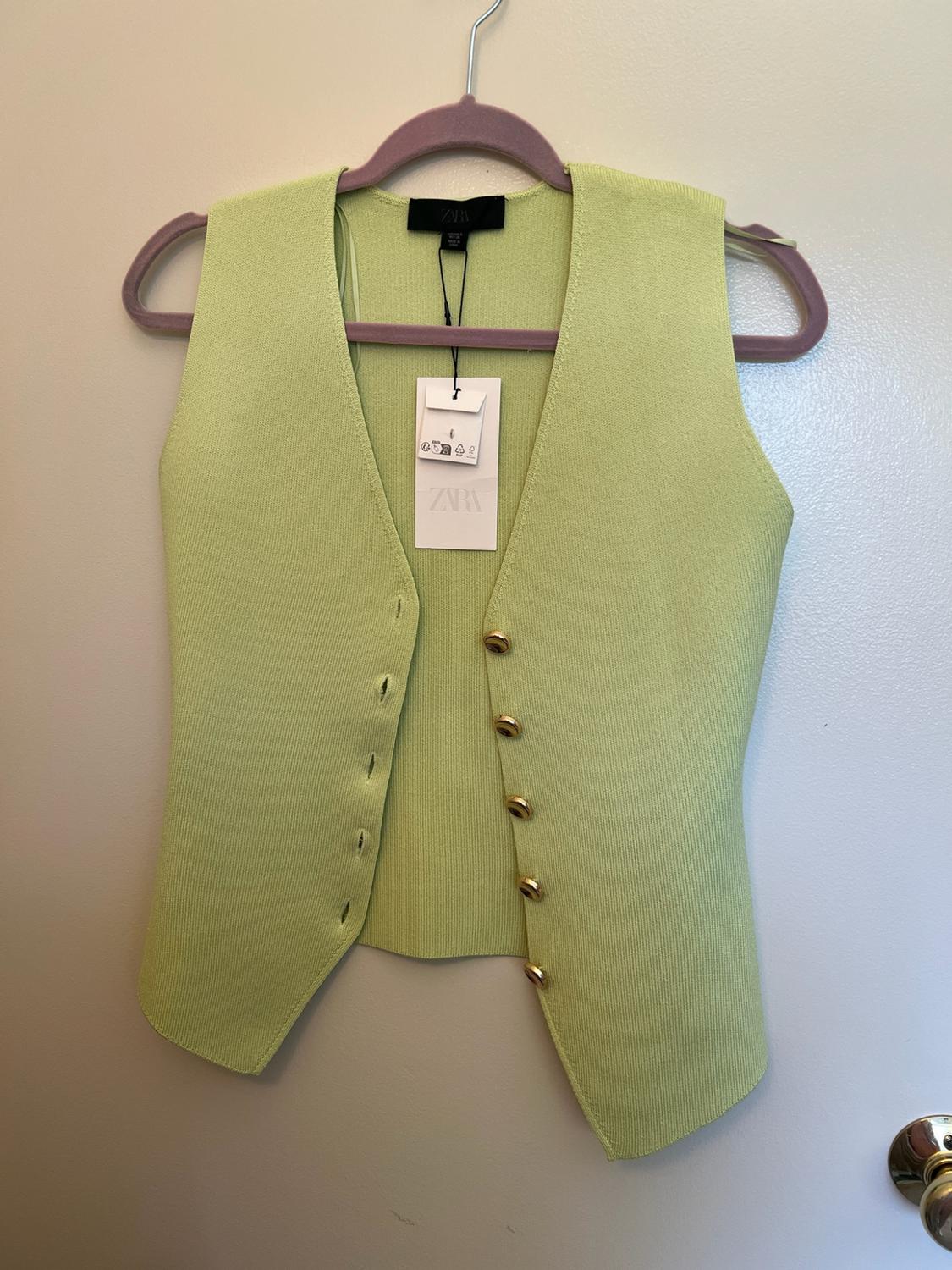 Light green knitted waistcoat with V-neck and gold buttons, hanging on a lilac hanger against a white wall. Label with text ZARA.