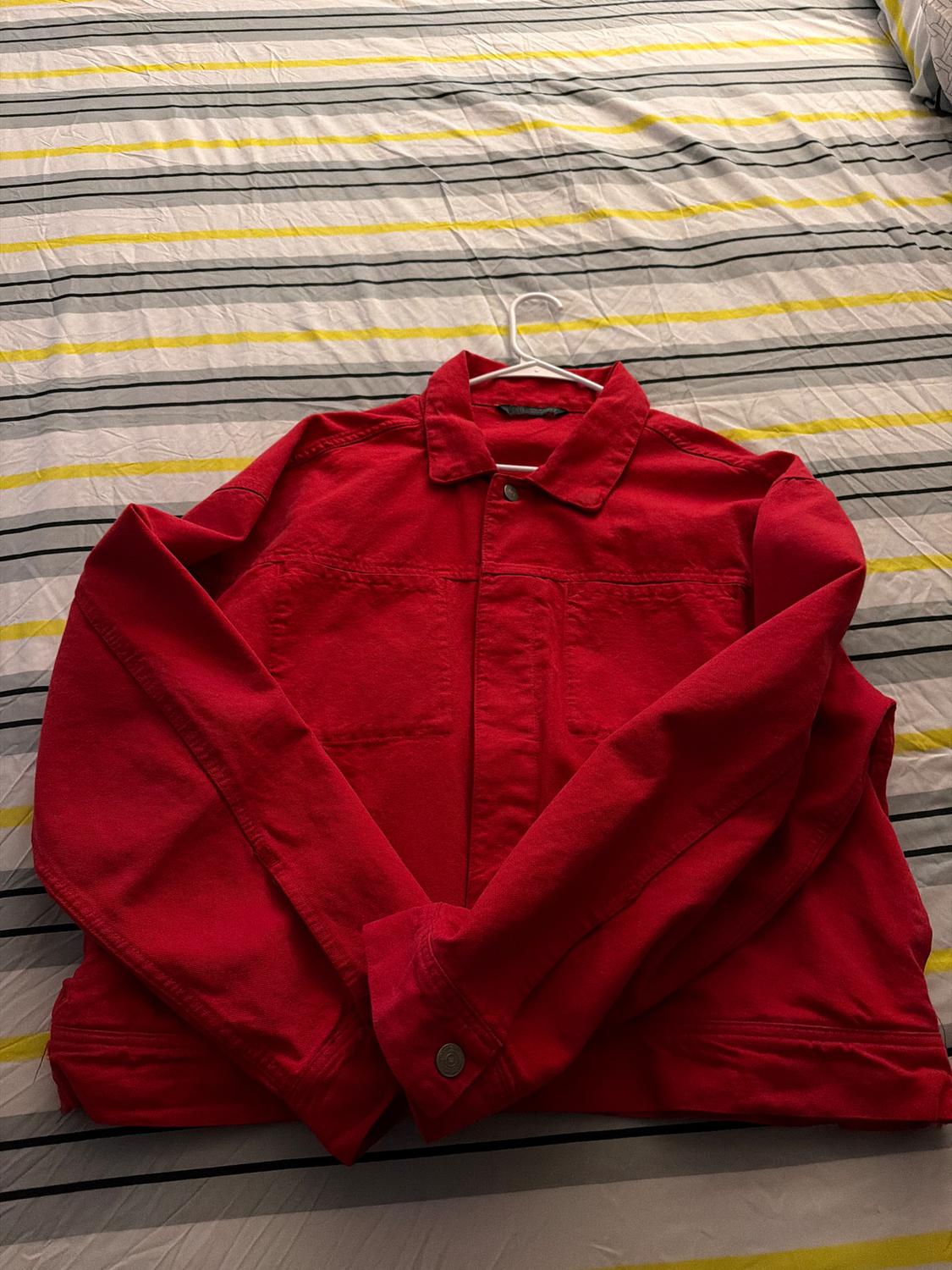 Red fabric jacket on a bed with a grey and yellow striped duvet. The jacket is on a white hanger.