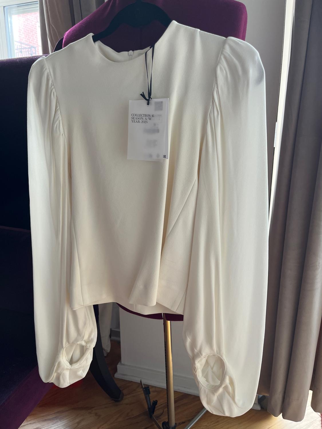 White long-sleeved blouse with a tag that reads "COLLECTION 16 SEASON A/W YEAR 2024", hanging on a black hanger over a purple chair.