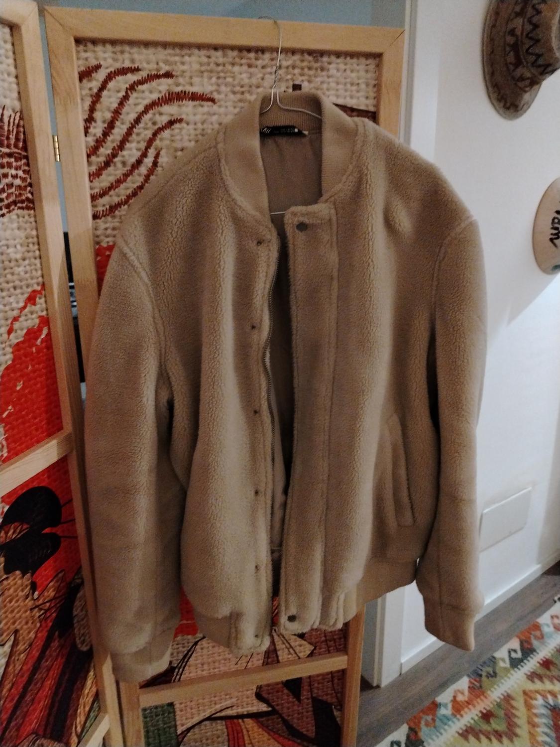Beige wool jacket hanging on a wooden coat rack, with a decorative panel background and a colourful rug on the floor.