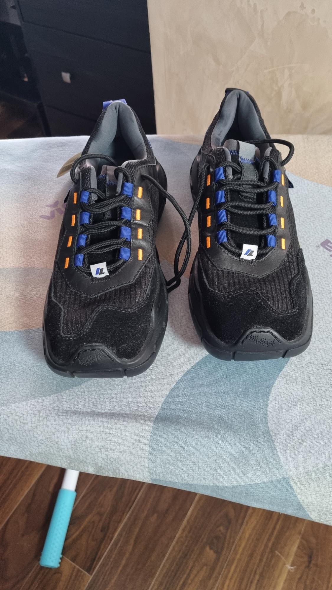 Black trainers with blue and orange details on an ironing board. Chunky soles and black laces. Wooden background and light-coloured wall.