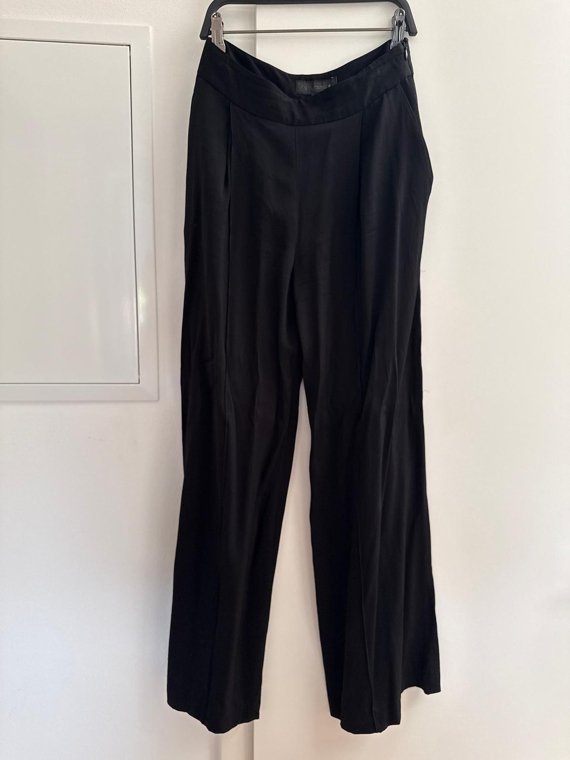 Black lightweight fabric trousers hung on a hanger against a white wall.