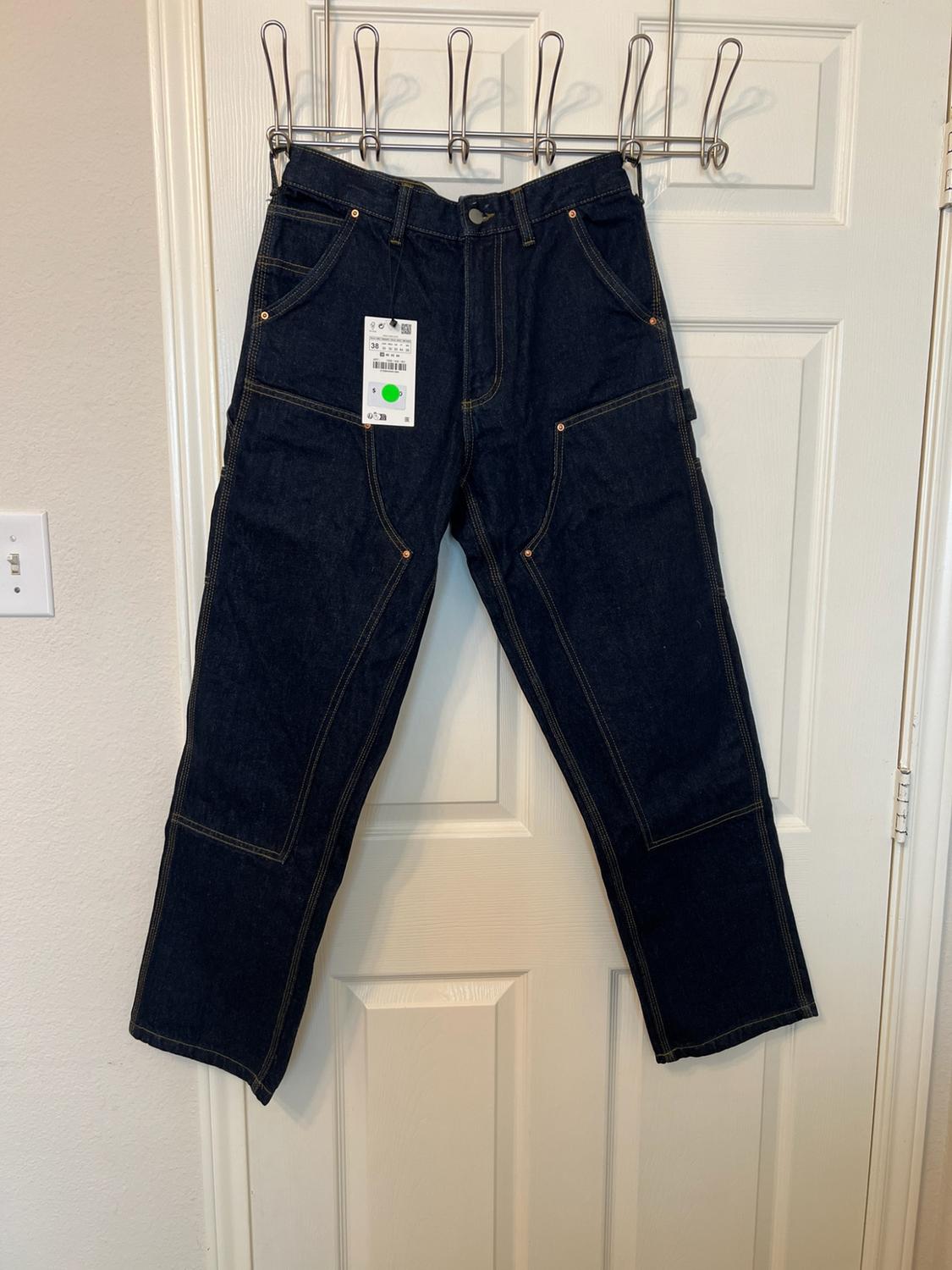 Dark blue denim trousers with contrasting stitching and front panels, hanging on a white door.