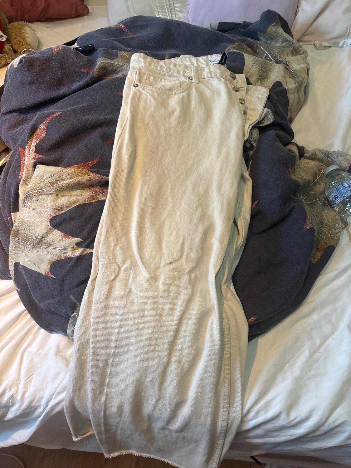 Light beige jeans with ZARA label, buttons and frayed hem, on an unmade bed with a patterned blanket and a water bottle.