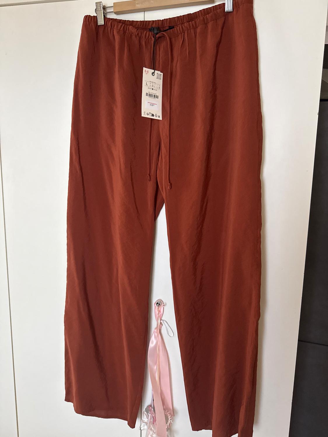 Terracotta wide-leg trousers hanging on a hanger, with a visible tag and decorative pink ribbons in the background.