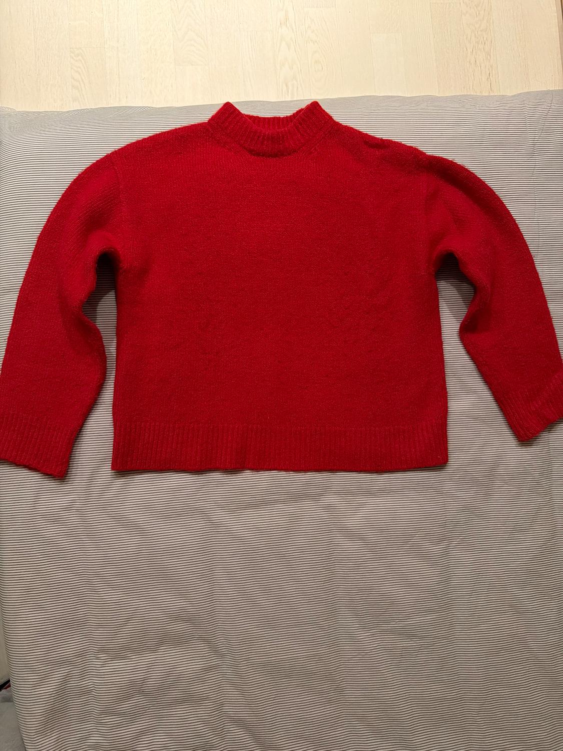Red knitted round-neck long-sleeved jumper, laid out on a grey and white striped fabric surface.