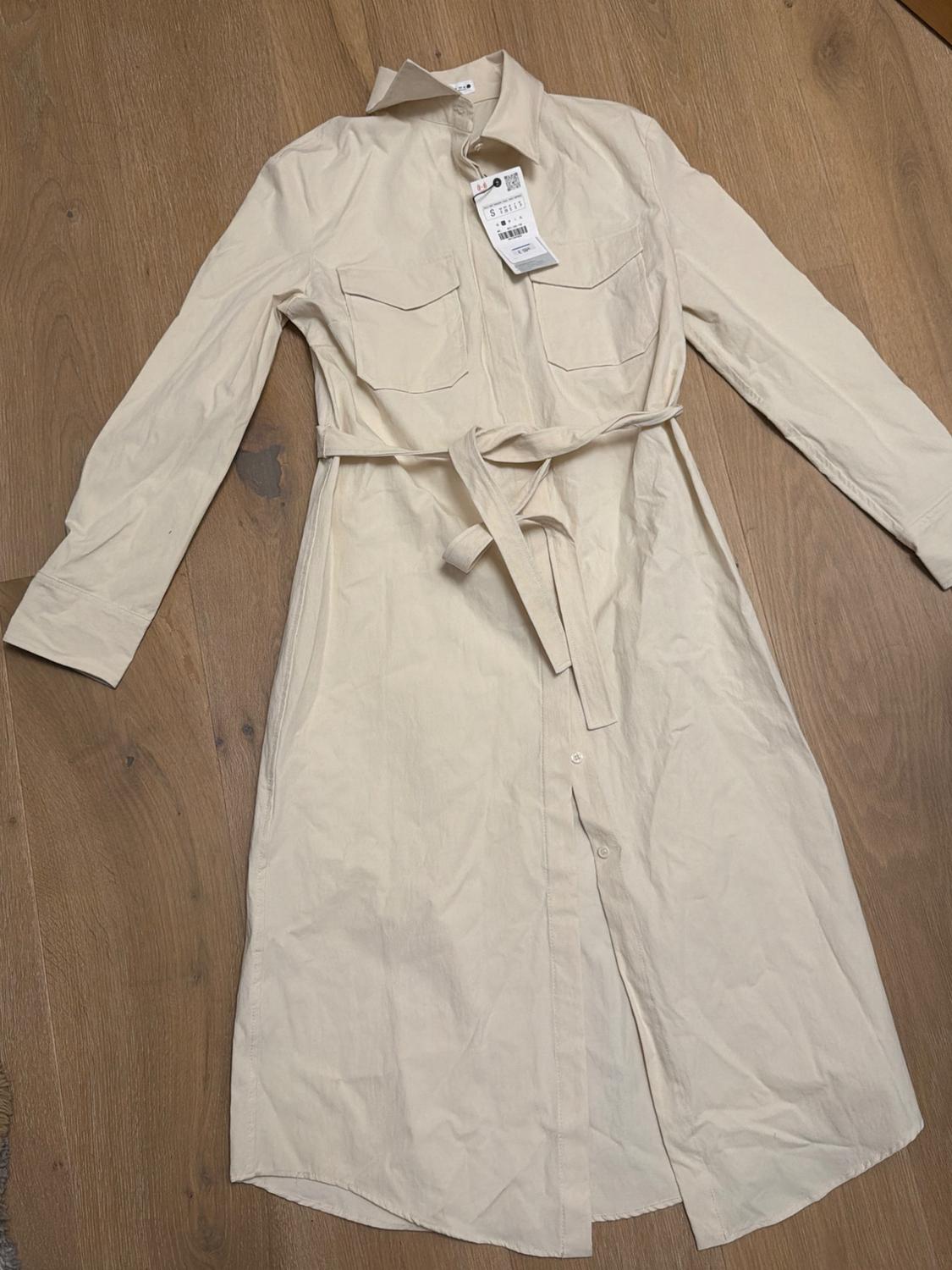 Beige long-sleeved dress with belt and front pockets on a wooden floor. Visible label with barcode.