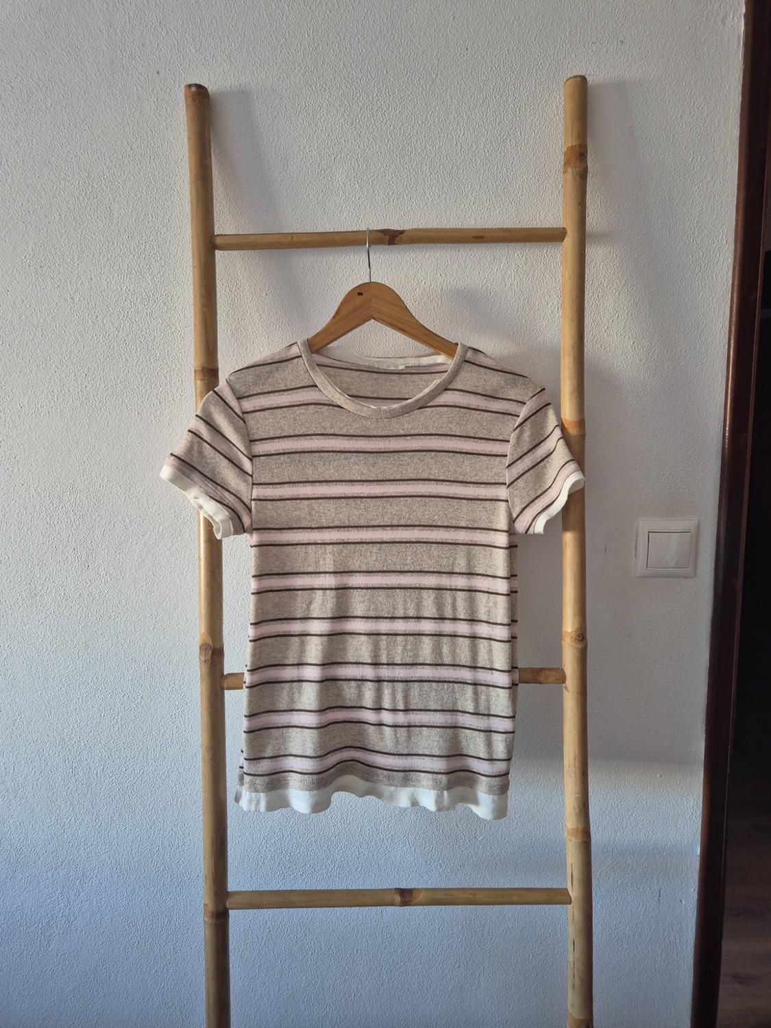 Beige, pink and brown striped short-sleeve t-shirt, hanging on a hanger on a bamboo ladder leaning against a white wall.