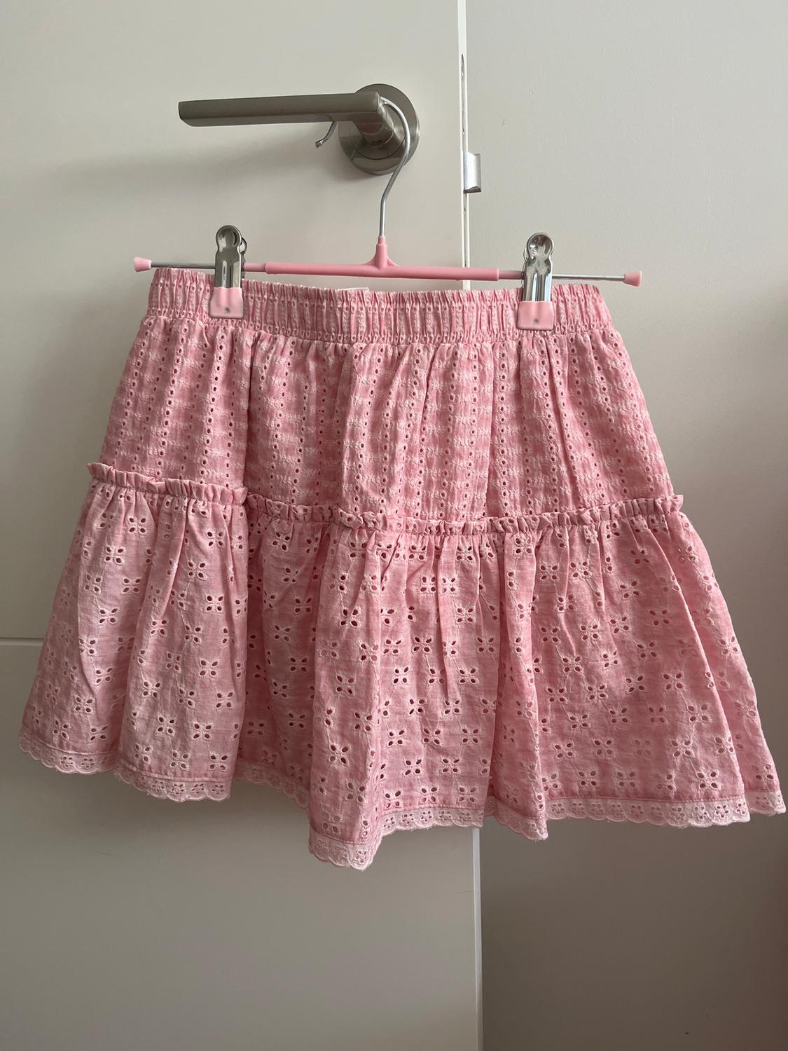 Pink skirt with floral embroidery hanging on a pink hanger over a white door with a silver handle.