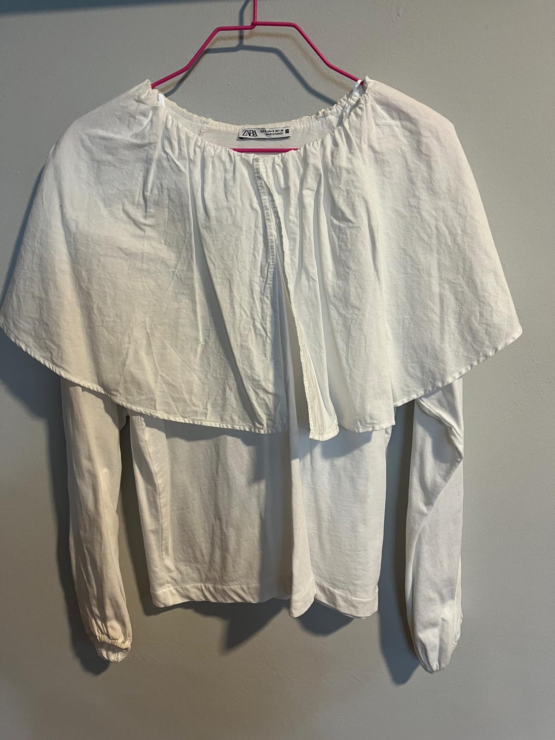White long-sleeved blouse with a wide collar, hanging on a pink hanger against a grey wall. Visible label: "Zara".