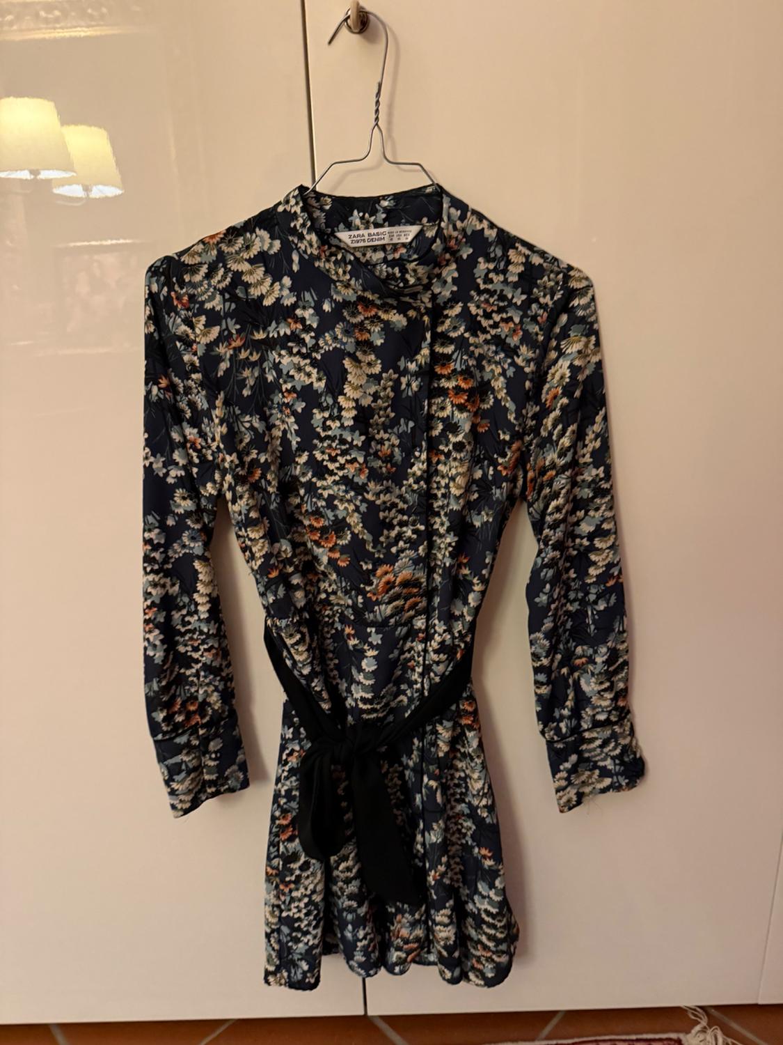 Floral long-sleeved dress hanging on a wire hanger, with a dark background and black belt, over a white door.