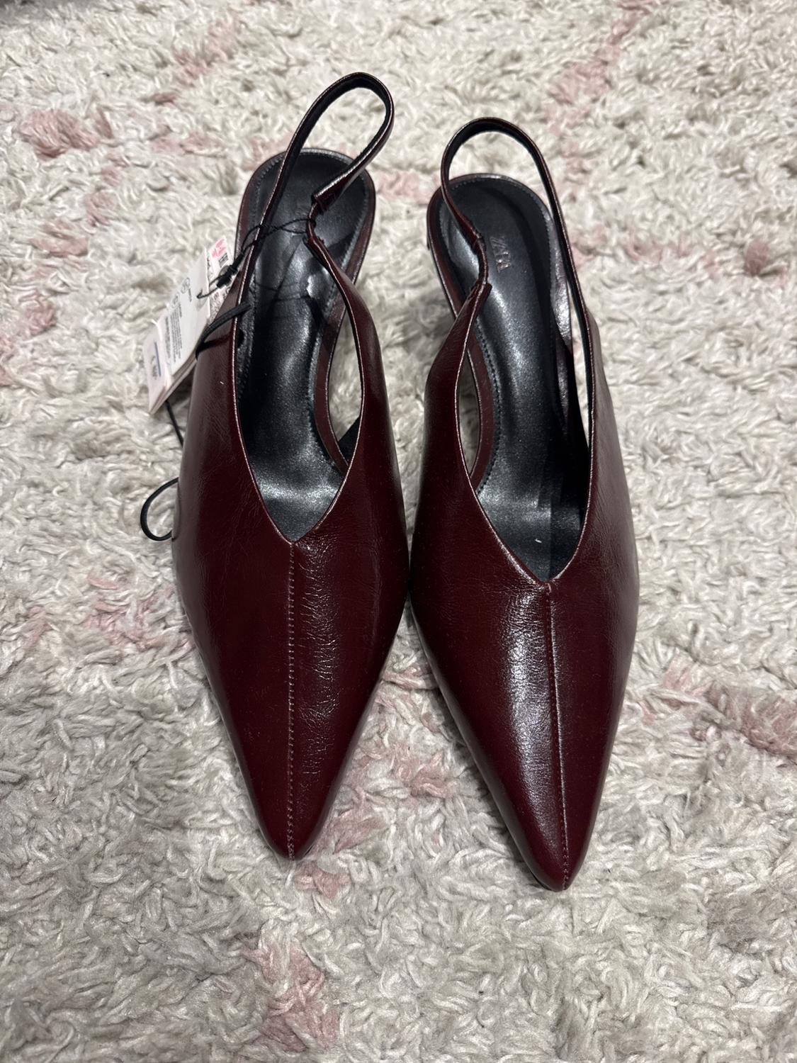 Burgundy high heels with a pointed toe and slingback straps, on a grey rug with pink details. Visible label on one shoe.