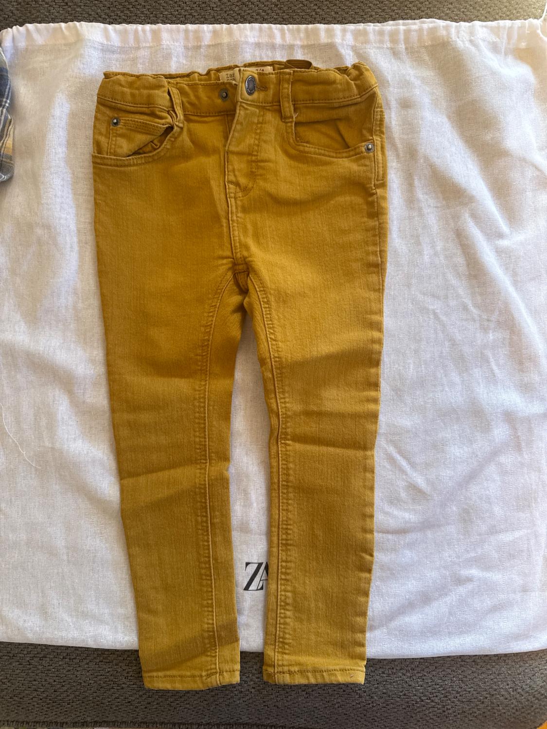 Yellow children's trousers on white fabric, with front pockets and a metal button. Part of a checked shirt visible on the left.