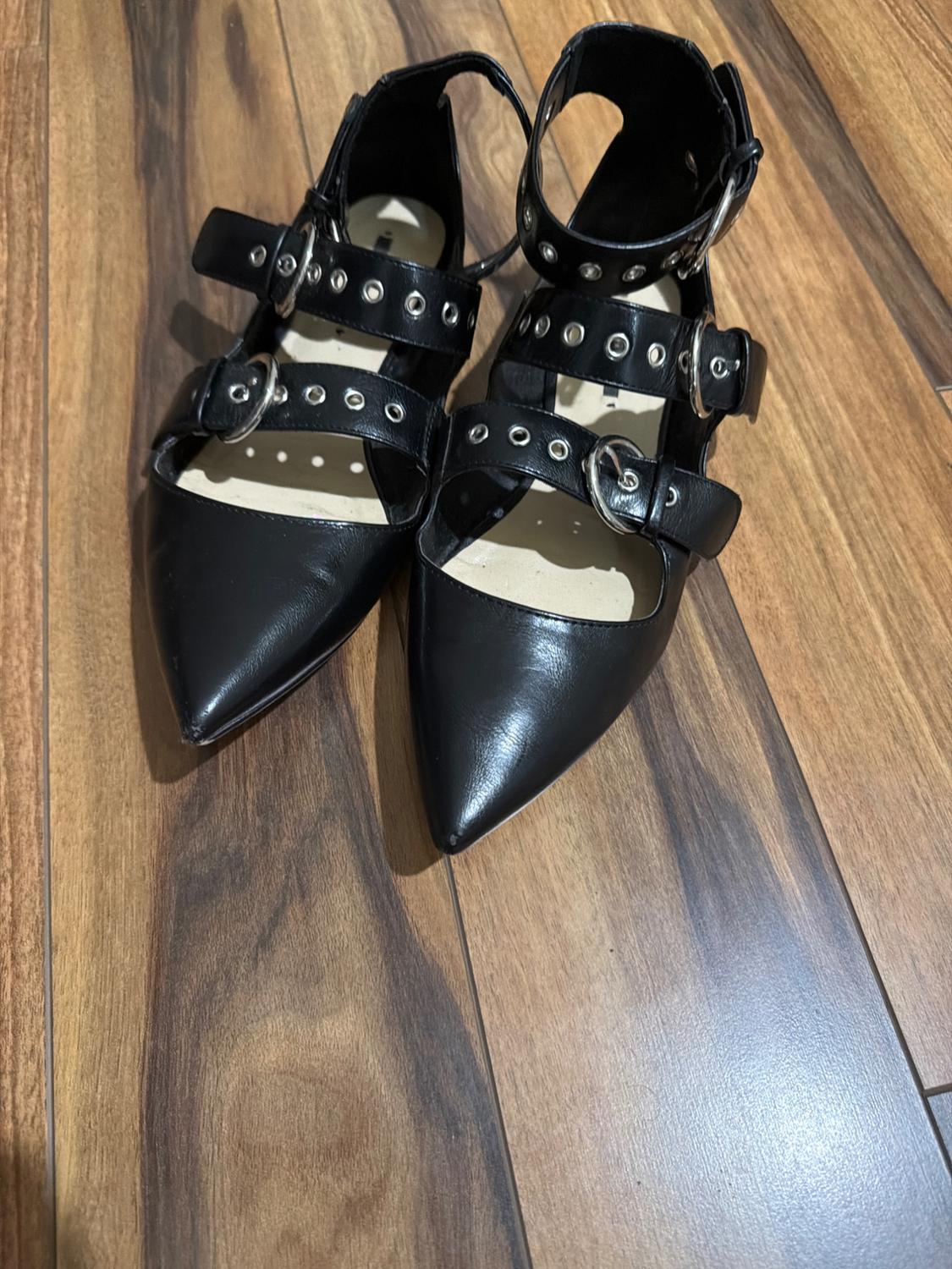 Pair of black pointed-toe flat shoes with multiple straps, buckles, and silver metallic details on a wooden floor.