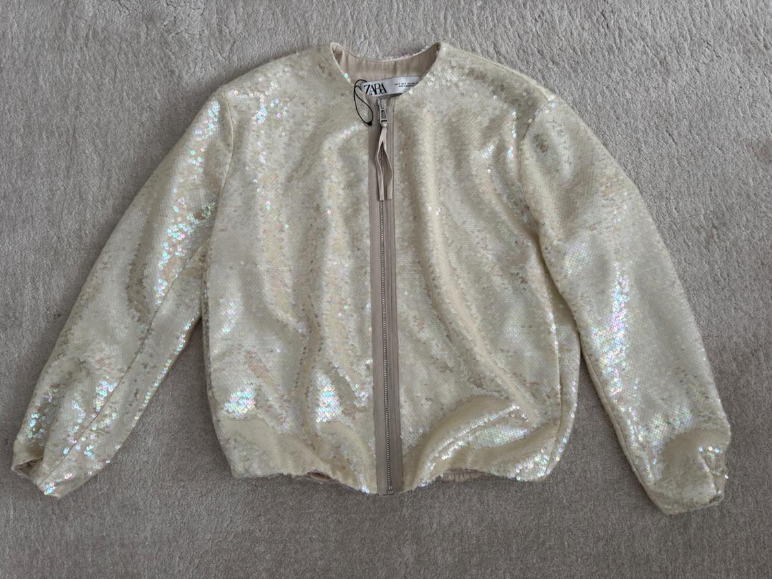 Cream iridescent sequin jacket with front zip, on a grey background. Visible label with text "ZARA".