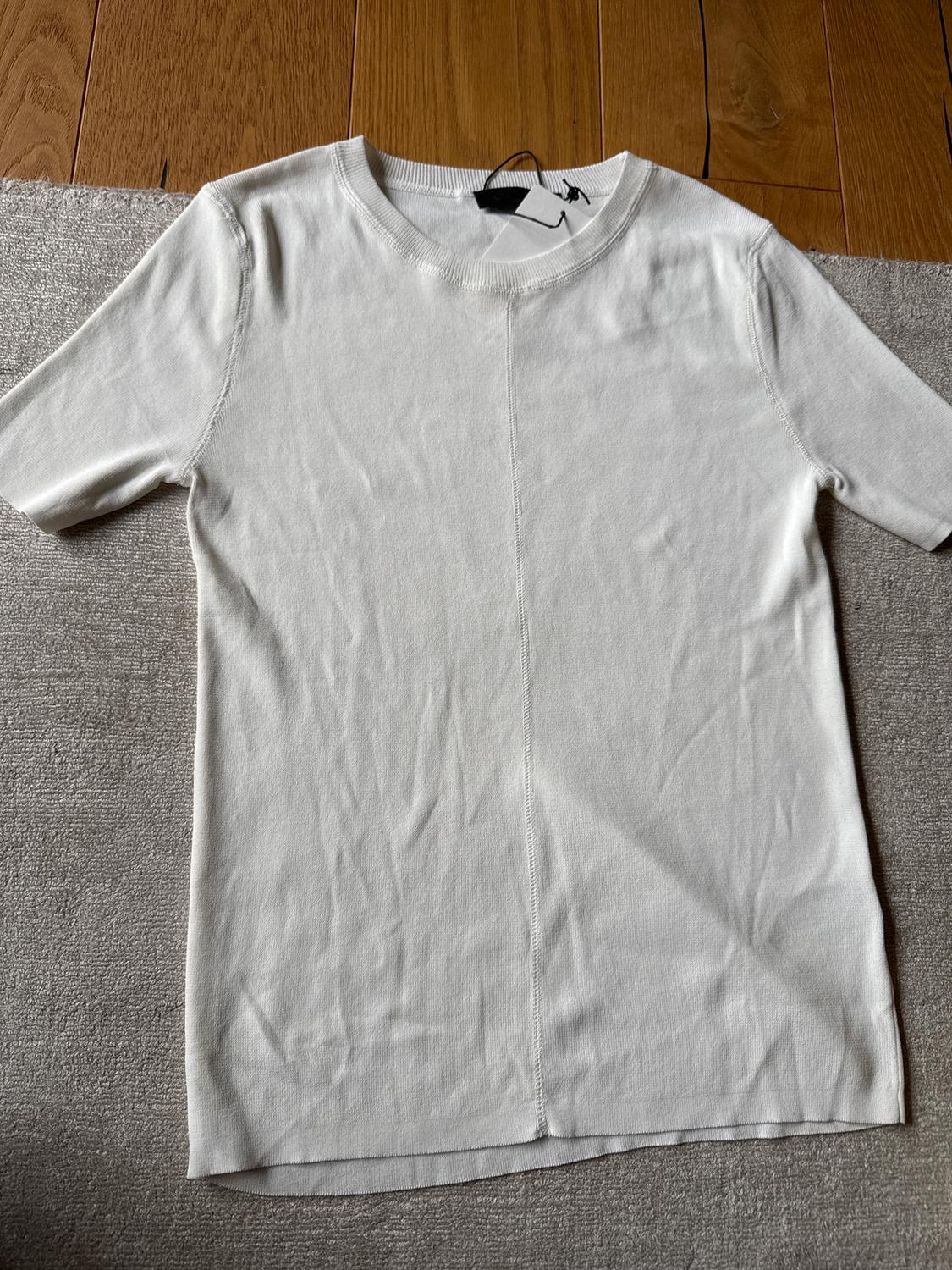 White short-sleeved t-shirt on a grey rug and wooden floor. Black tag visible on the collar.