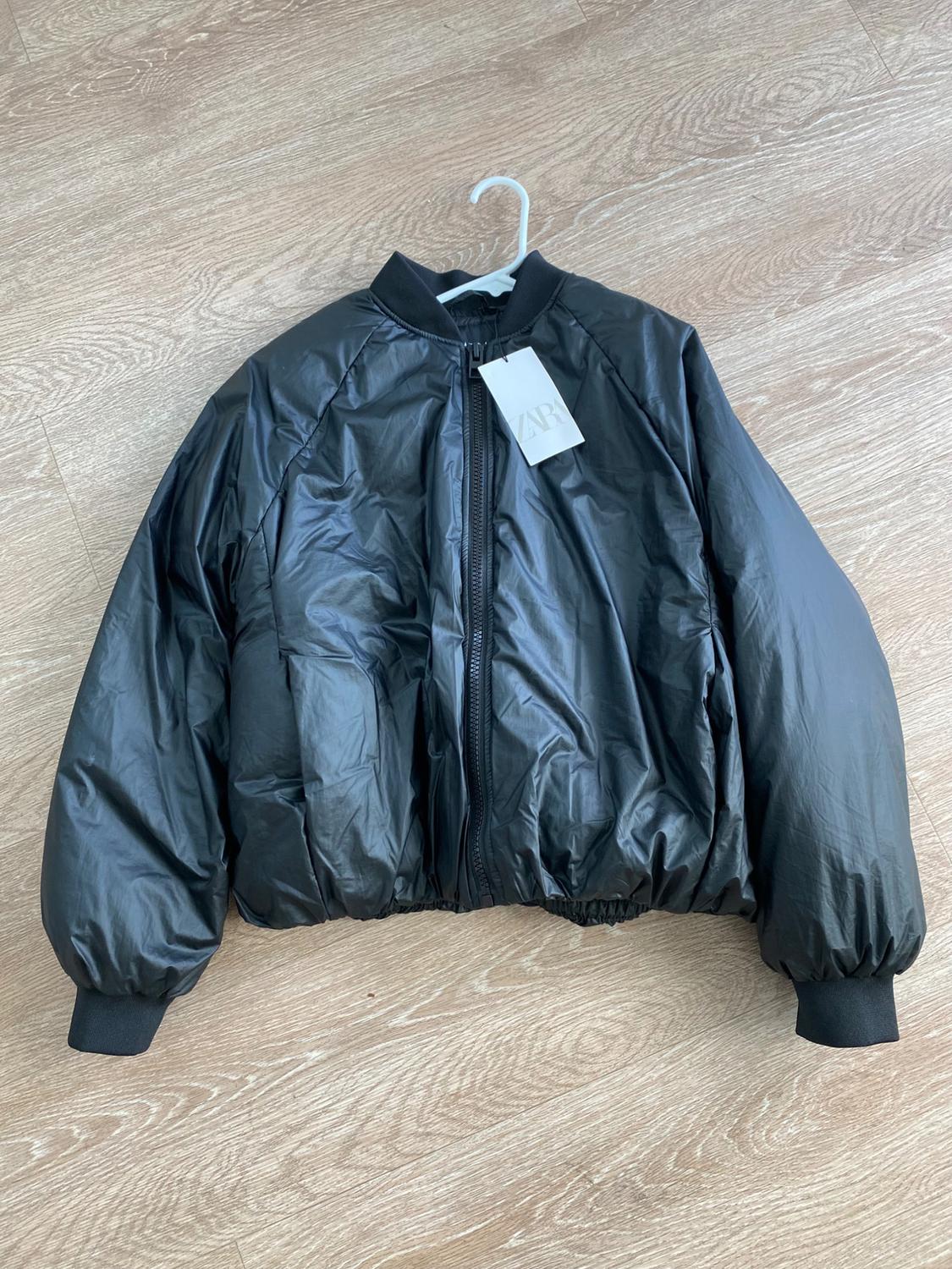 Black quilted jacket with zipper and elastic cuffs, hanging on a hanger over a light wood floor. Visible tag.