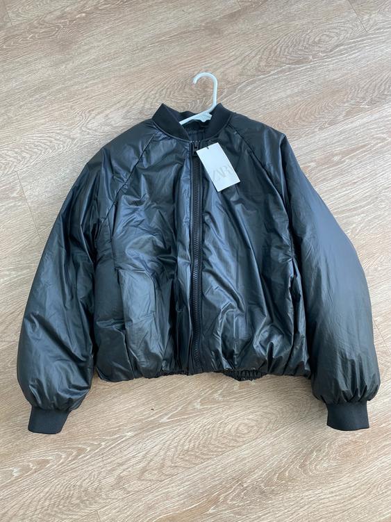 Black quilted jacket with zipper and elastic cuffs, hanging on a hanger over a light wood floor. Visible tag.