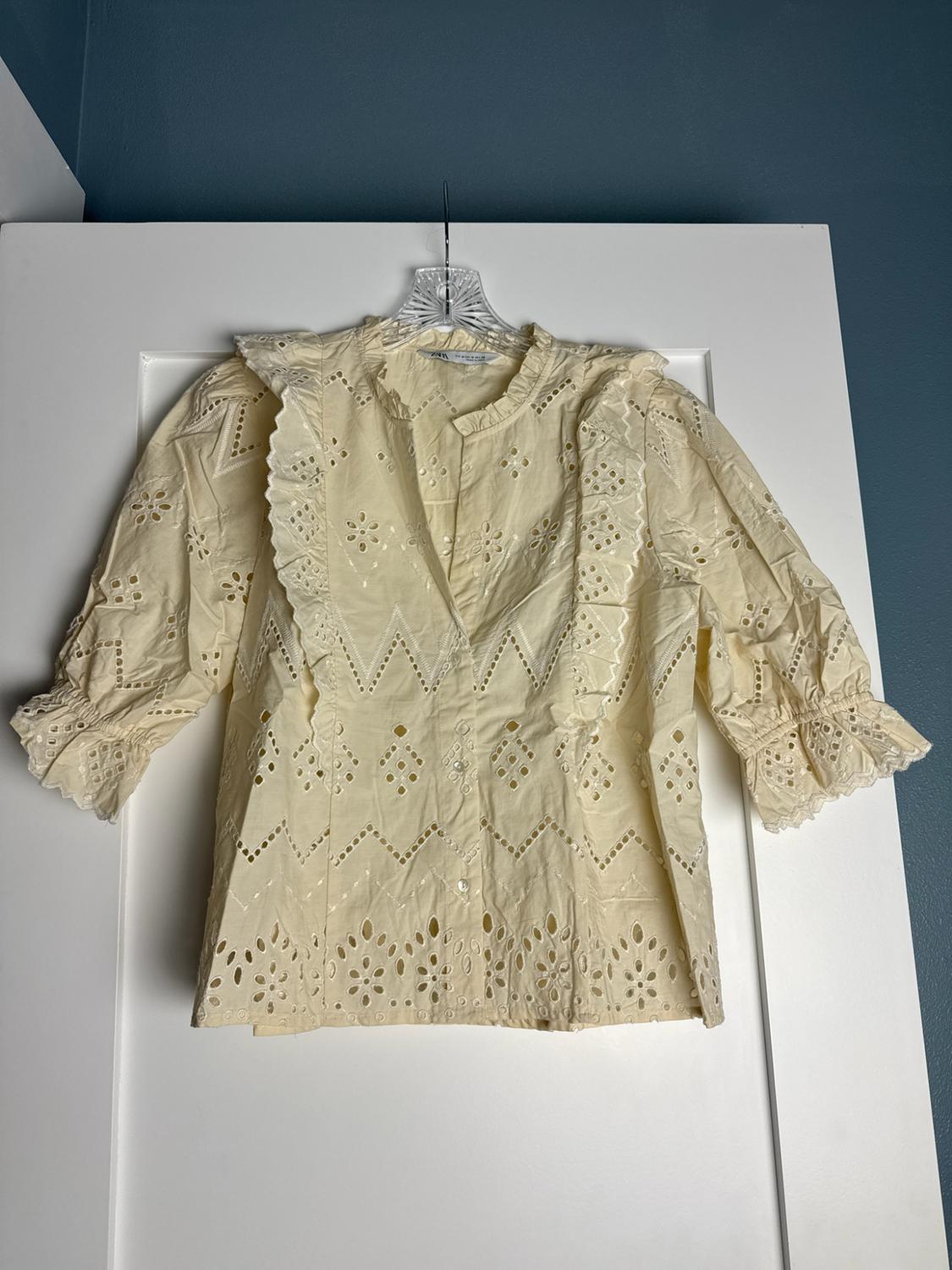 Cream blouse with broderie anglaise, front buttons and frill details, hanging from a hanger on a white door.