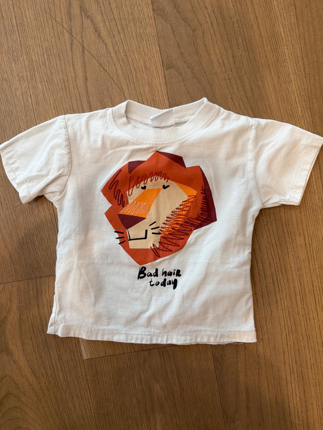 Children's white T-shirt with a lion drawing in orange tones and the text "Bad hair today" on a wooden floor.