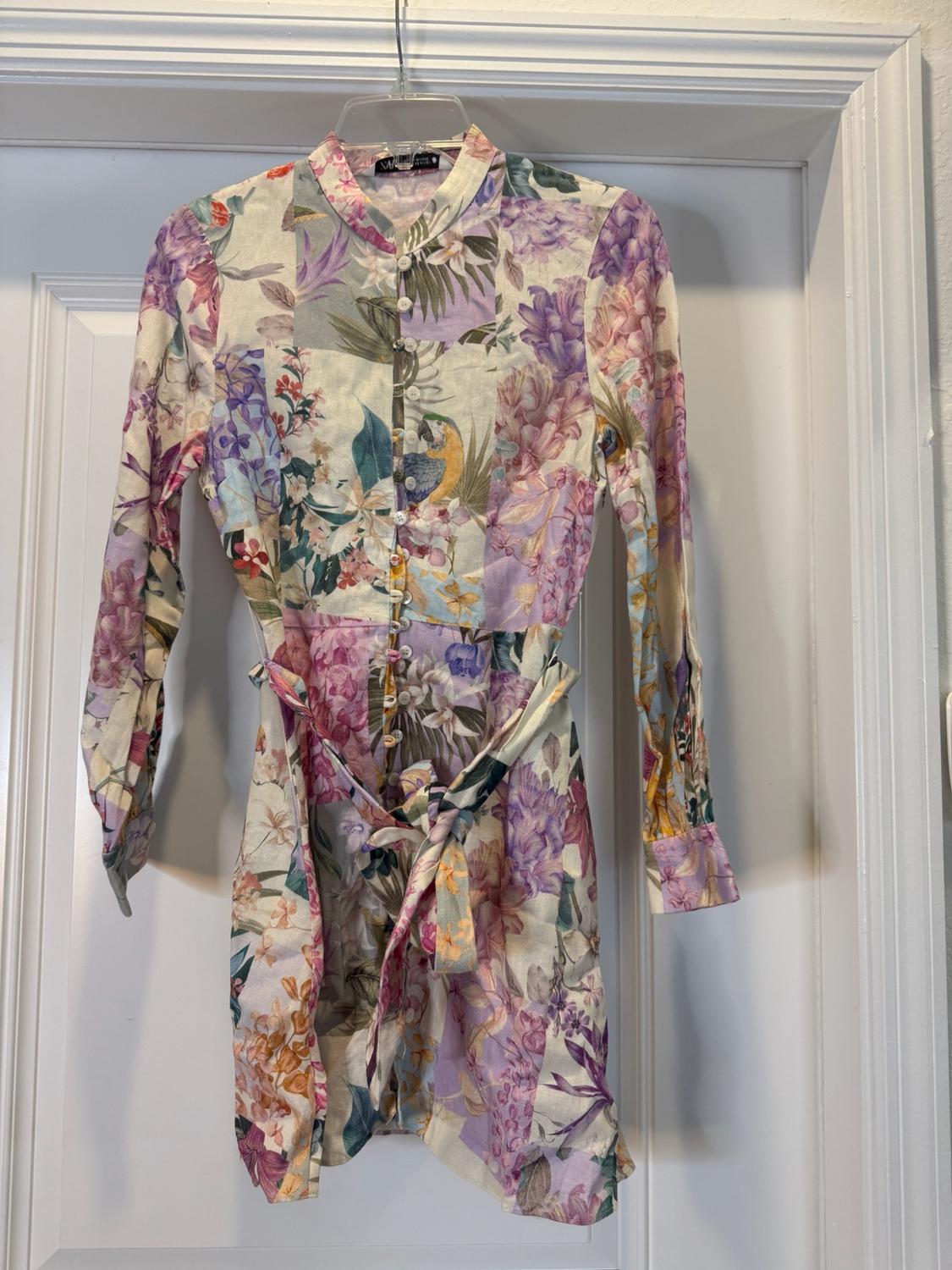 Pastel floral and parrot print long-sleeved dress with a tie belt, hanging on a hanger over a white door.