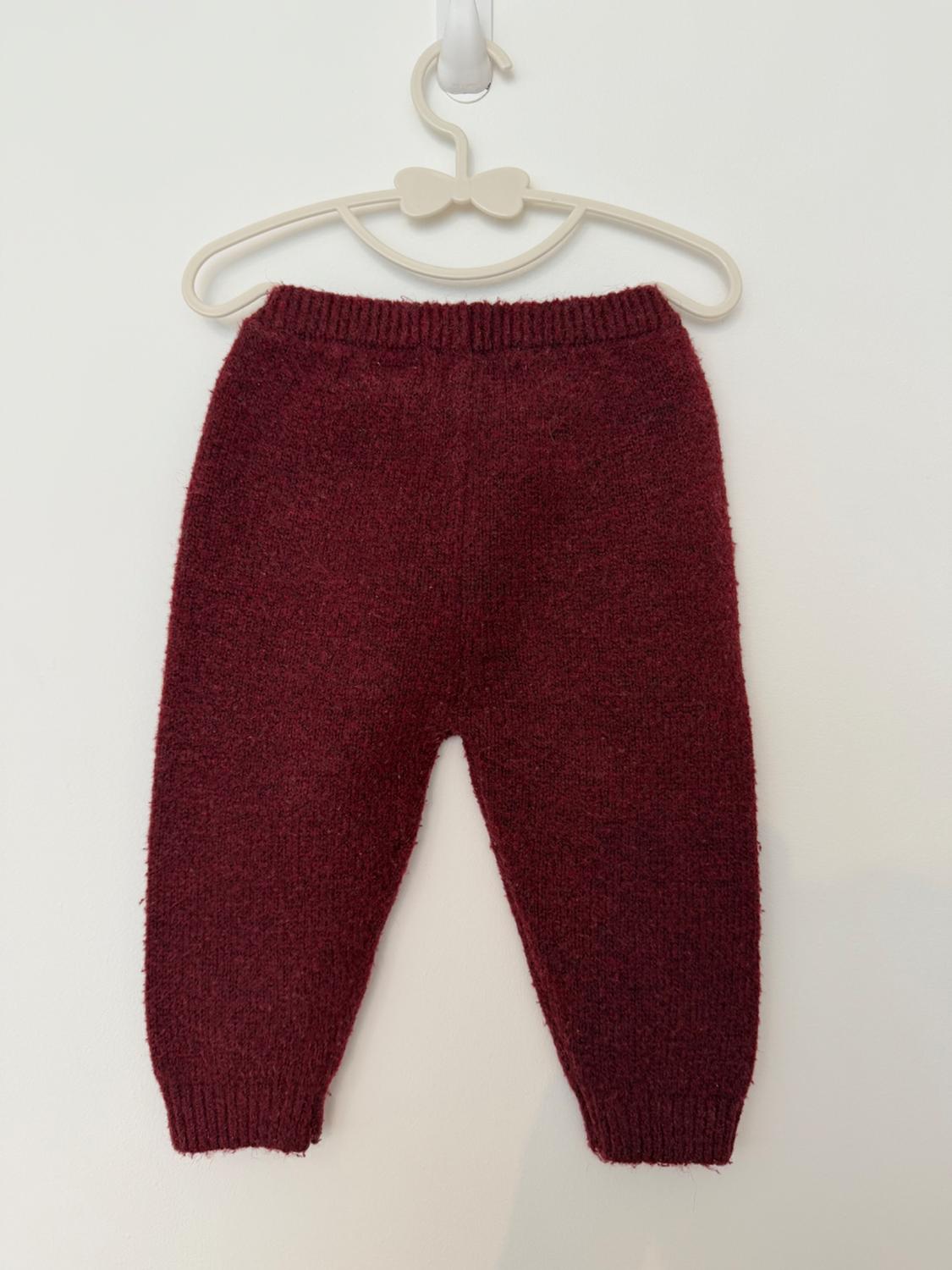 Maroon knit trousers hanging on a white bow-shaped hanger against a plain white background. Minimalist and cosy composition.