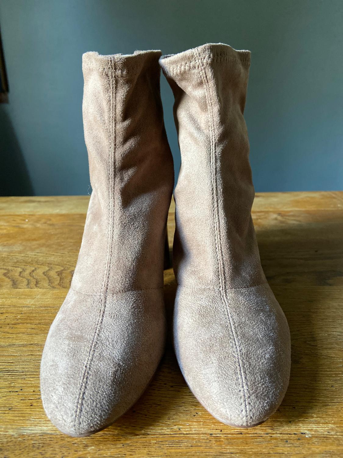 Pair of beige ankle boots with front stitching, resting on a wooden table
in front of a greyish-blue wall.