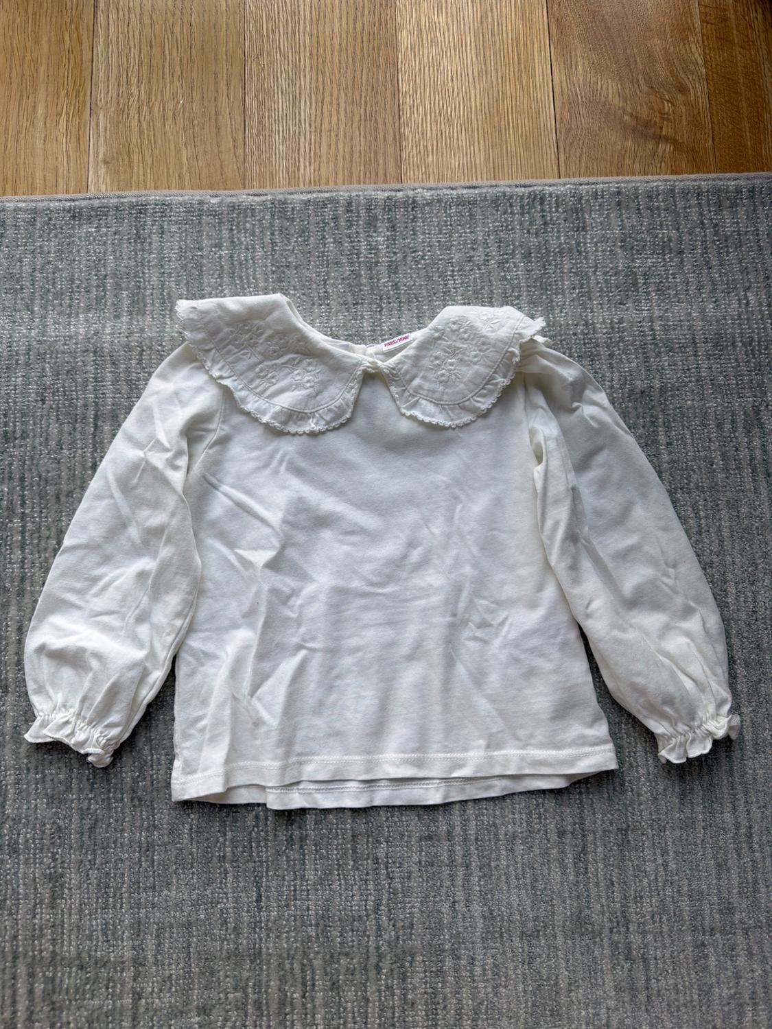 Long-sleeved white blouse with embroidered collar, on a grey rug and light wood flooring.