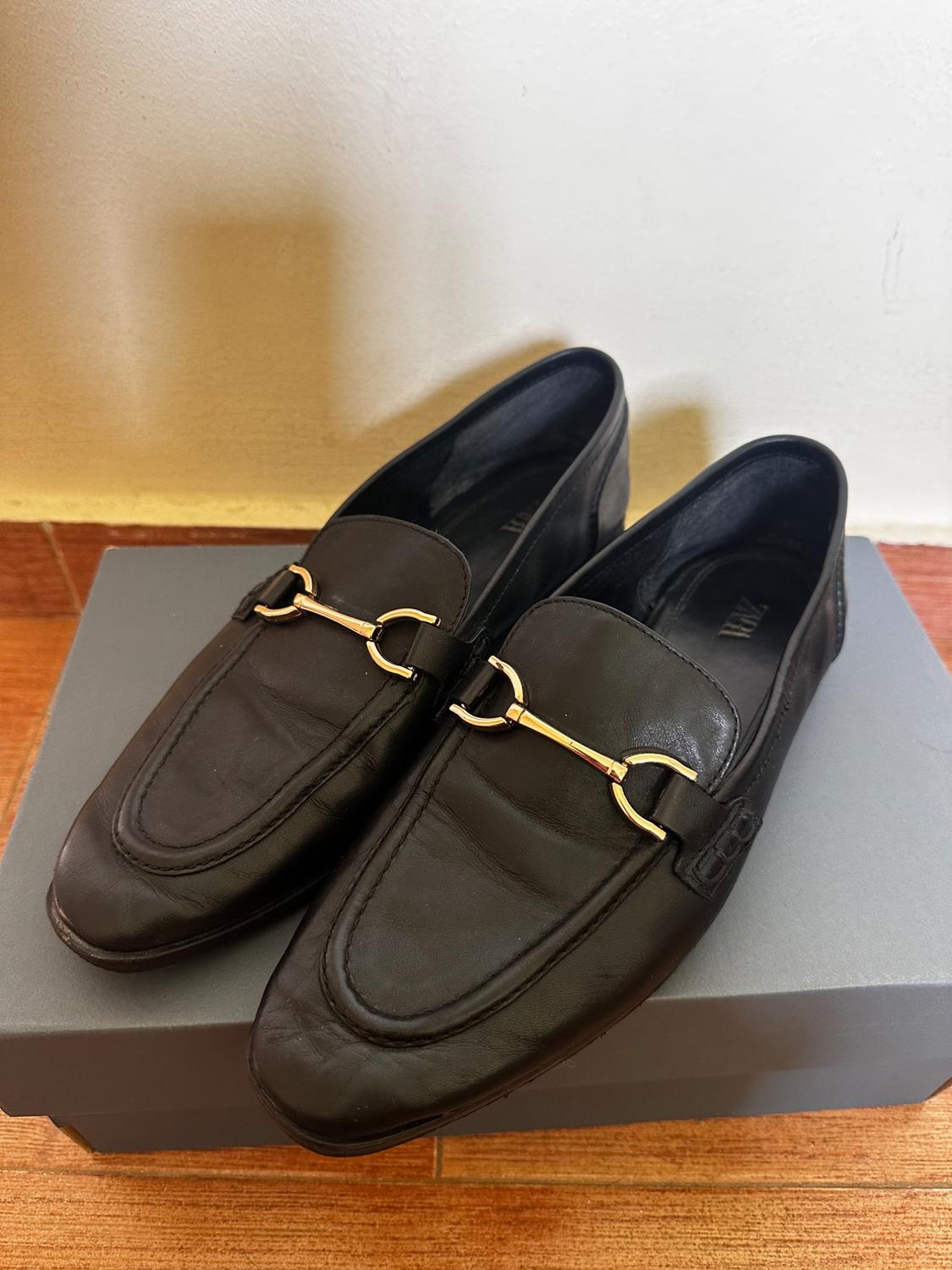 Black leather loafers with a gold metallic detail on the top, placed on a grey box on a wooden floor.